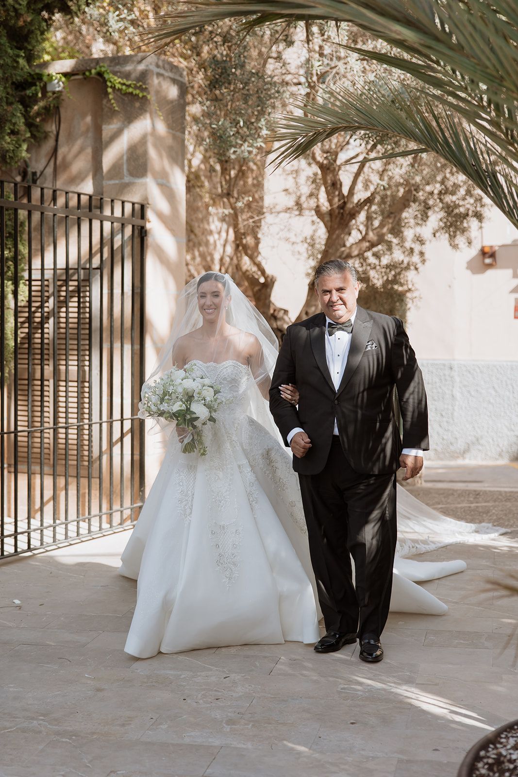 Couple walking along a palm-lined path during a formal wedding ceremony.