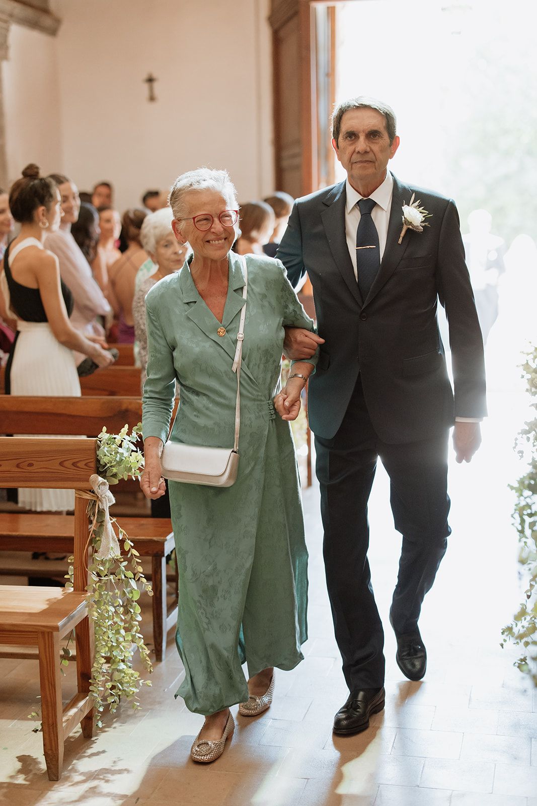 Elegant couple walks down church aisle, woman wearing sage green dress and man in black suit.