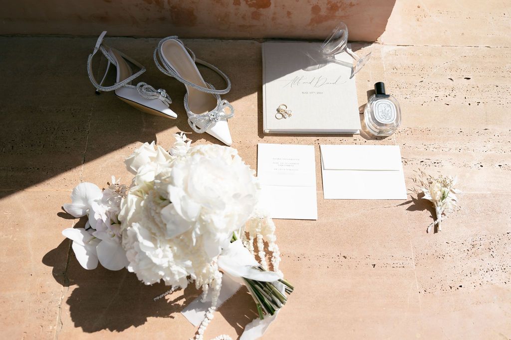 Wedding details flatlay with white floral bouquet, stationery and accessories in sunlight.