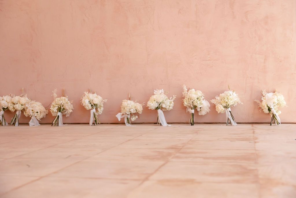 A row of identical white floral wedding bouquets lined up against a peachy pink wall.