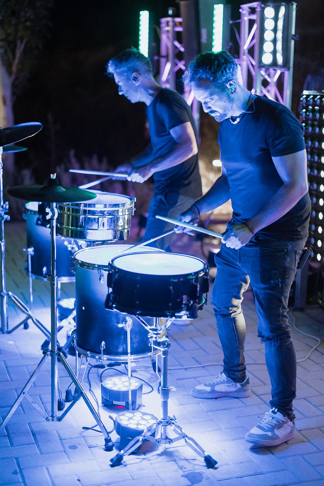 Two drummers performing with drums under dramatic blue stage lighting.