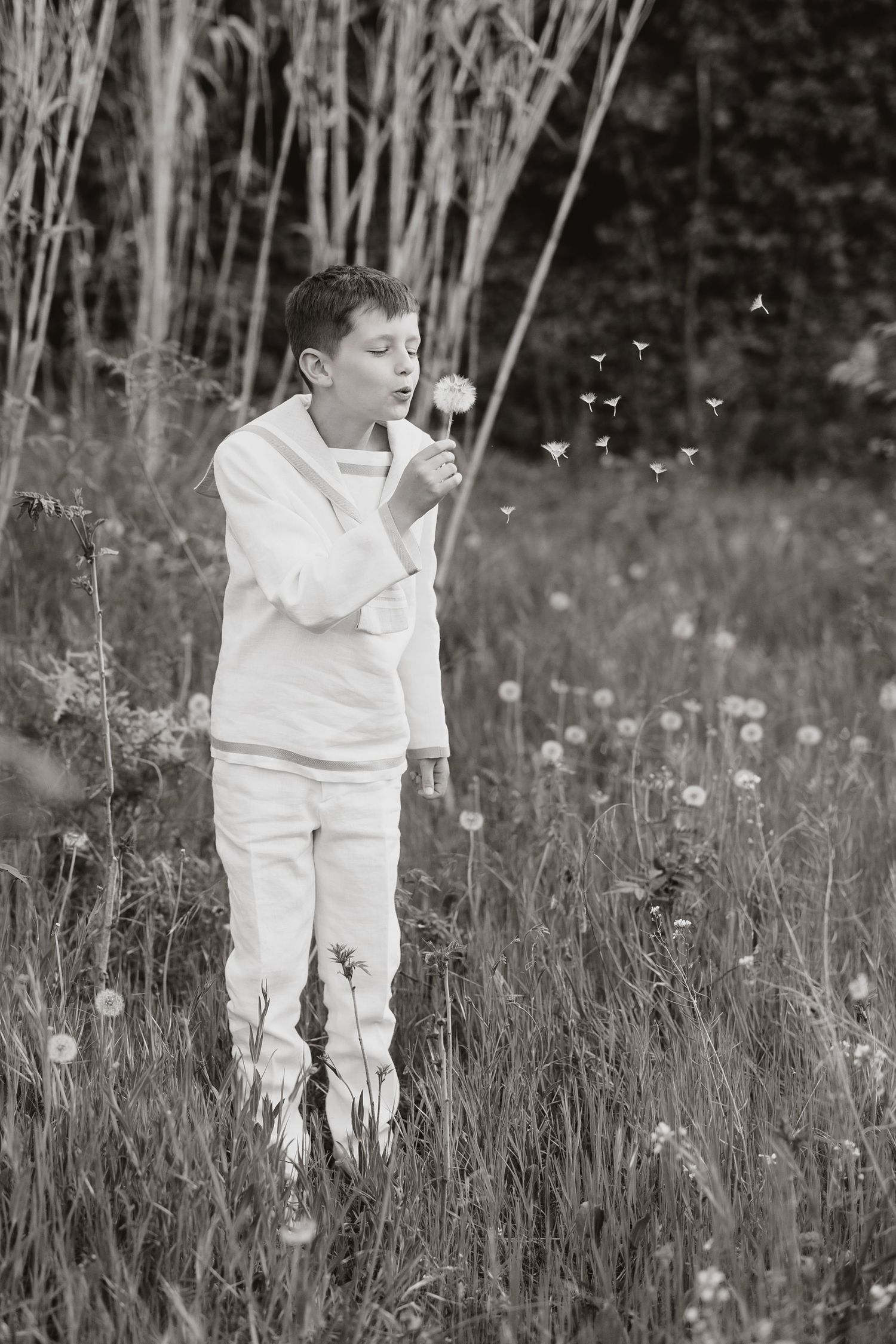 A young boy in white clothes blows dandelion seeds in a field, captured in black and white photography.