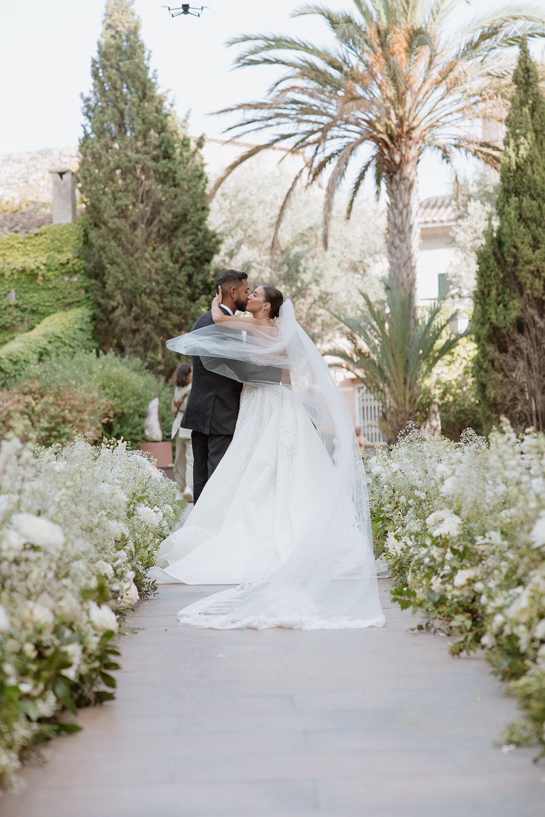 Wedding couple embracing in garden path lined with white flowers and palm trees.