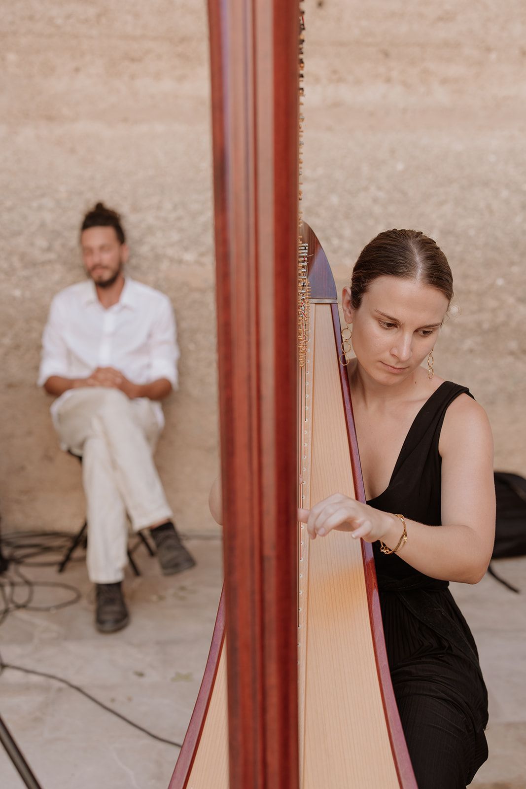 Professional harpist in black dress performing next to elegant wooden harp against stone wall.