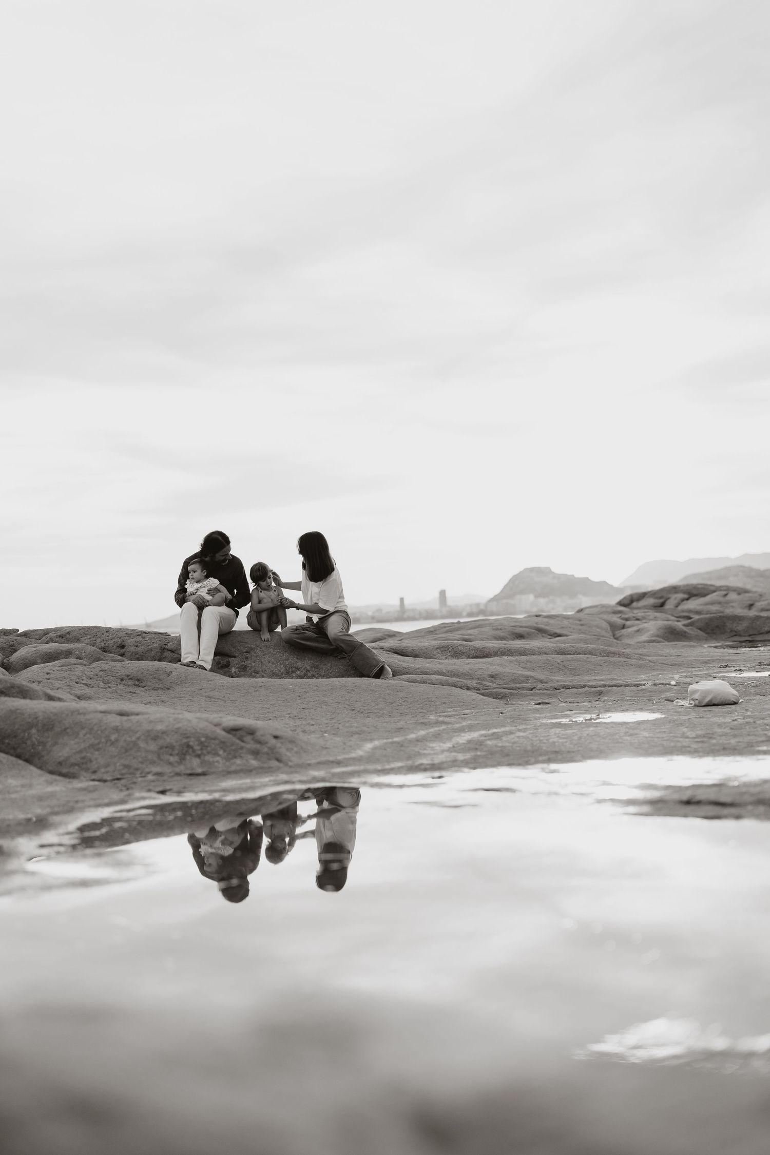Black and white image of figures on coastal rocks reflected in still water.
