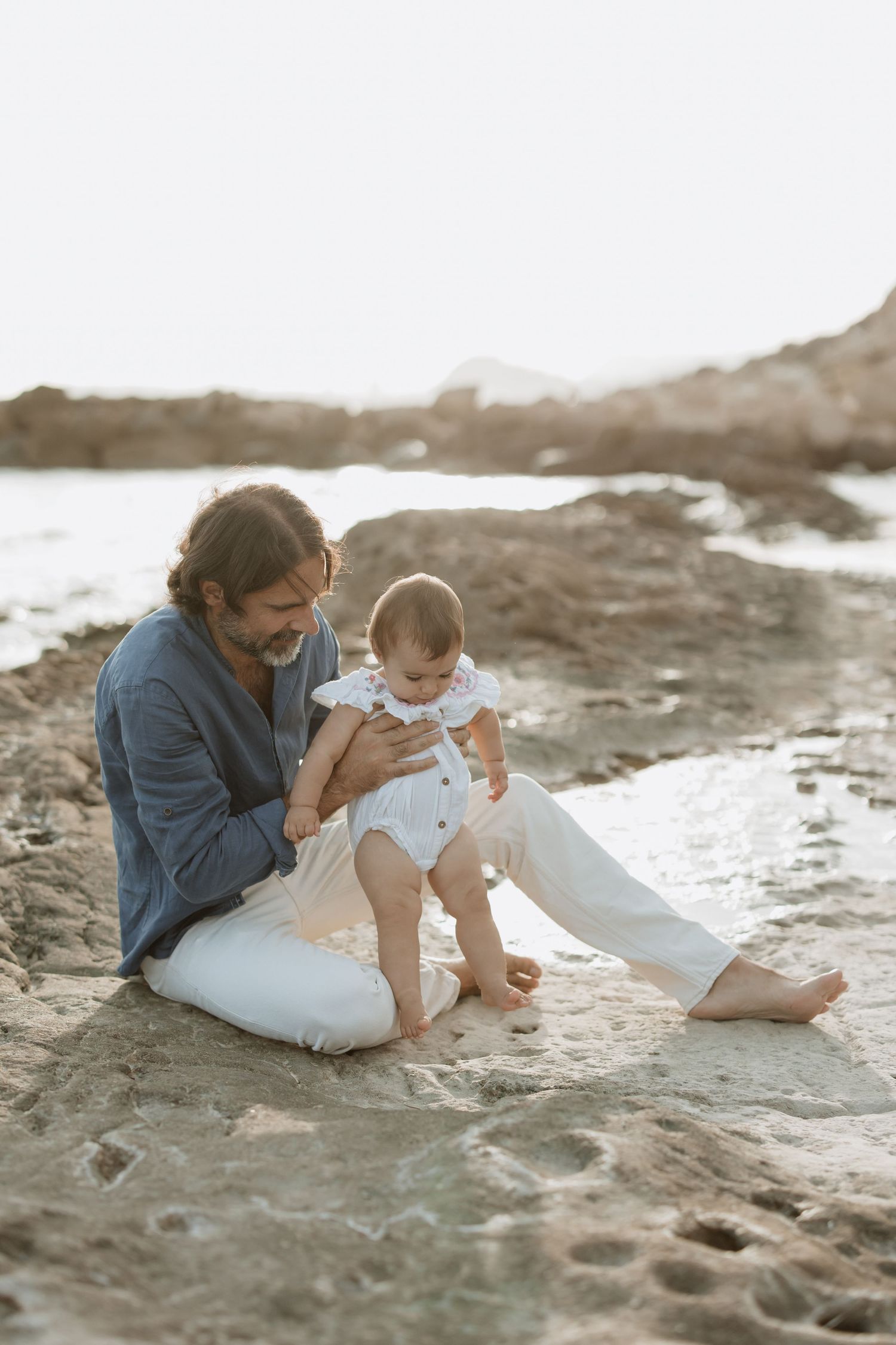 Relaxed moment between adult and baby sitting together on beach sand at golden hour.