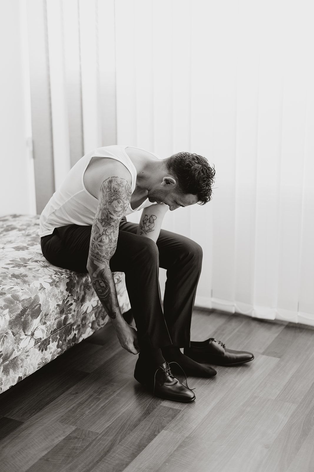 A groom in formal attire sits on a bed while getting ready.