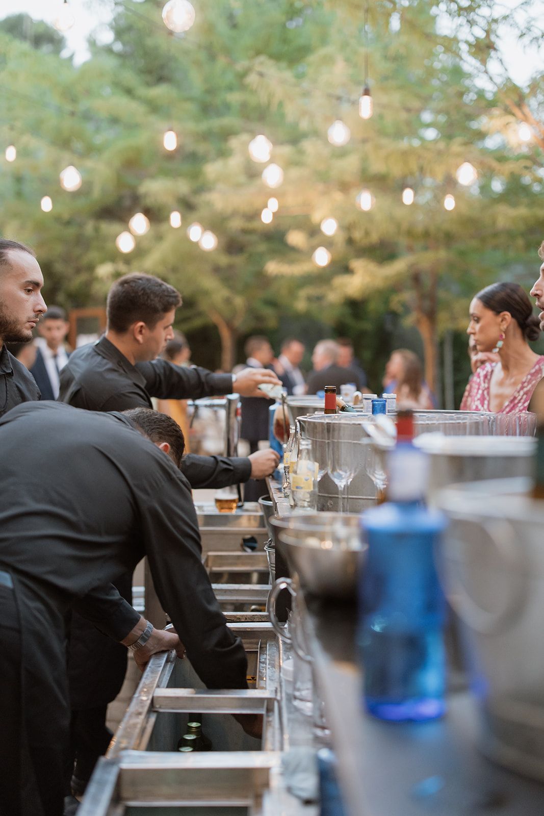 Bartenders setting up outdoor bar service station with string lights in background.