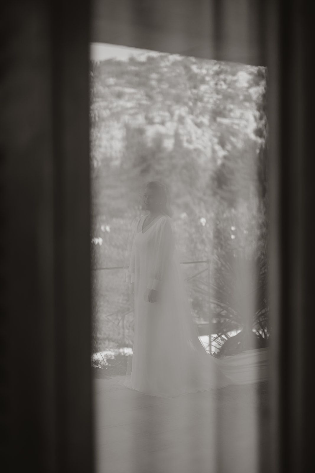 Dreamy silhouette of bride seen through window with soft garden backdrop.