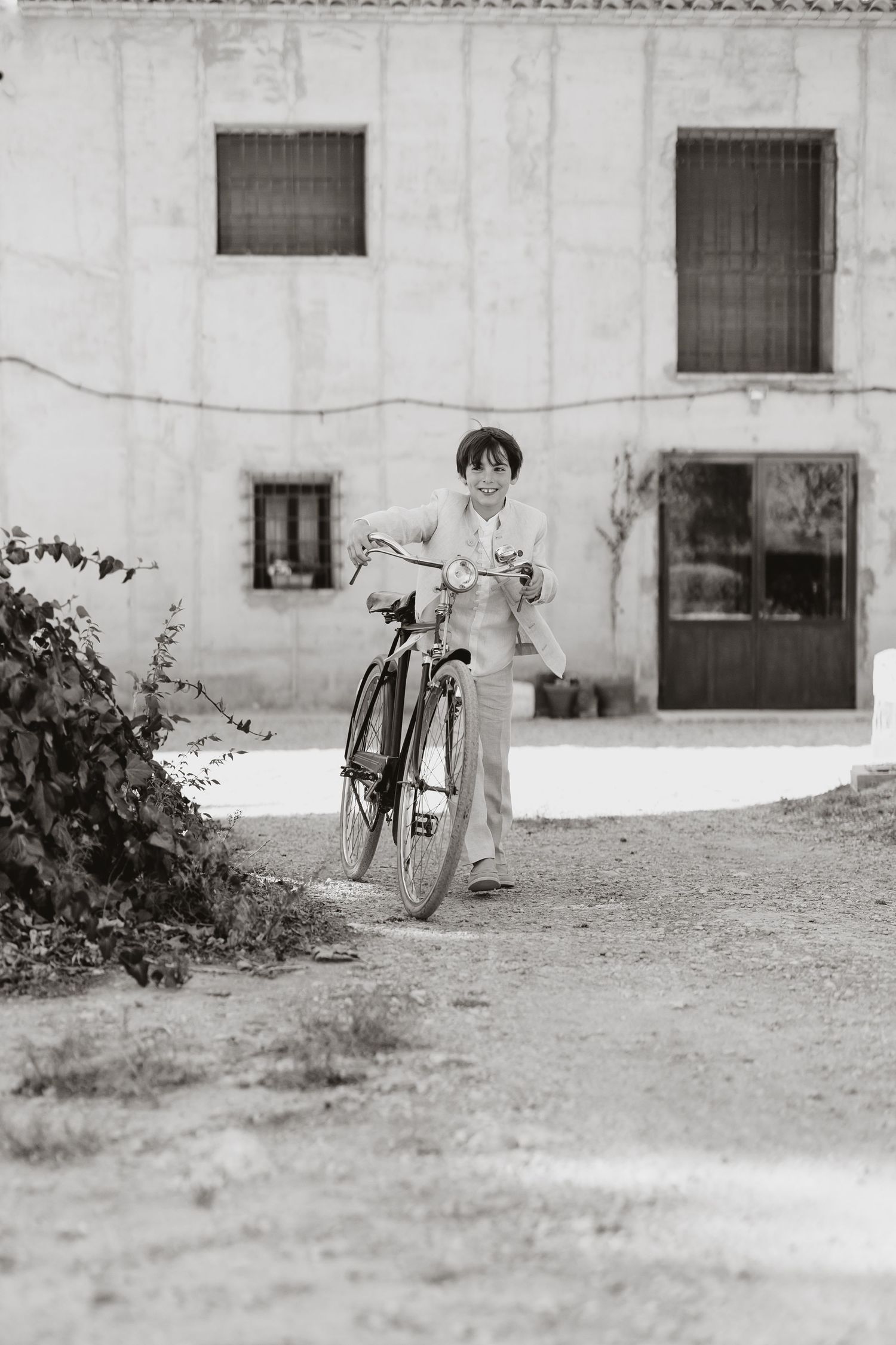 Black and white vintage photo of a small figure with a bicycle standing outside a white building with small windows.