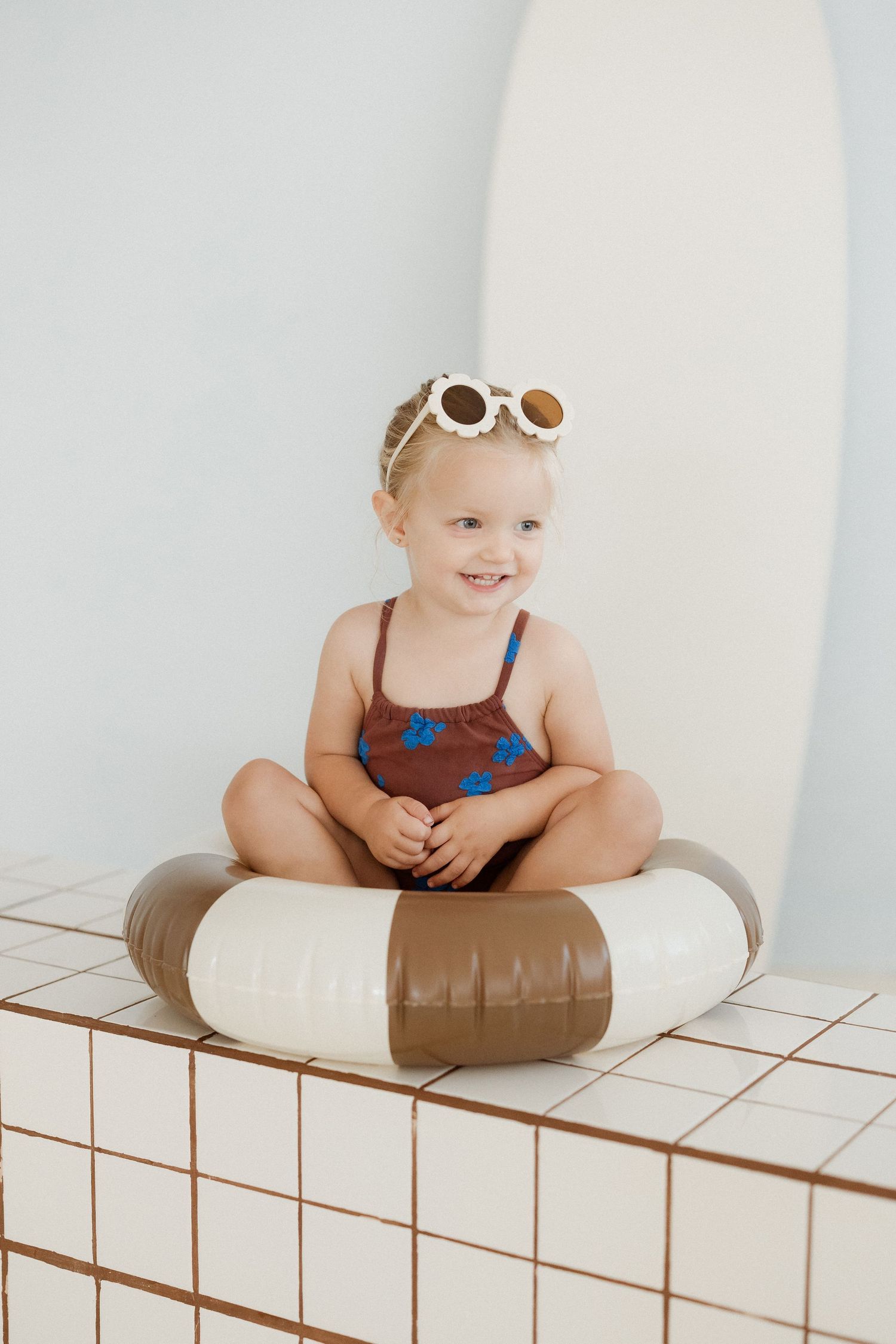Smiling toddler girl in brown swimsuit and sunglasses sitting in inflatable pool ring on tiled surface.