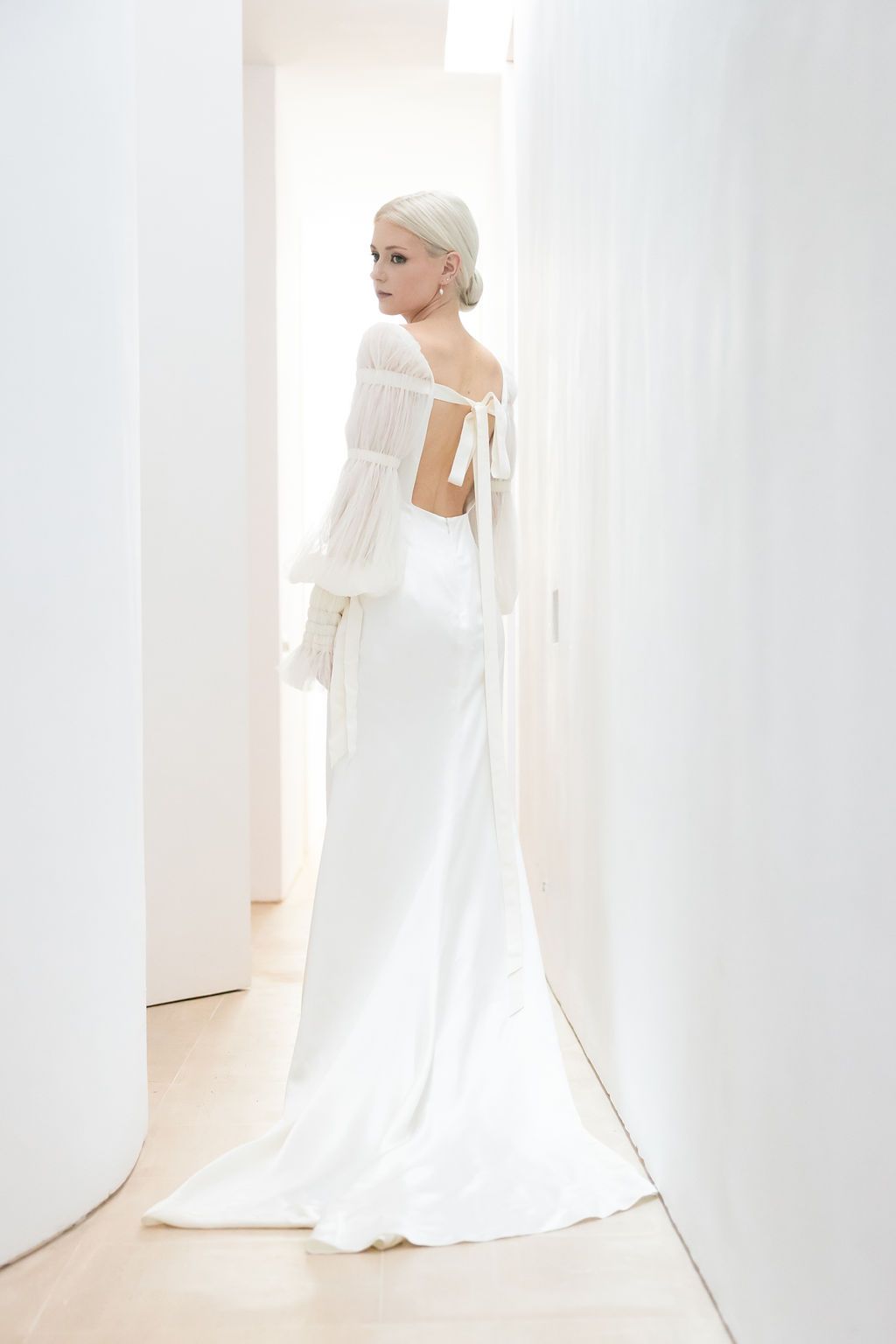 An elegant backless wedding gown with flowing train photographed in a minimalist white hallway.