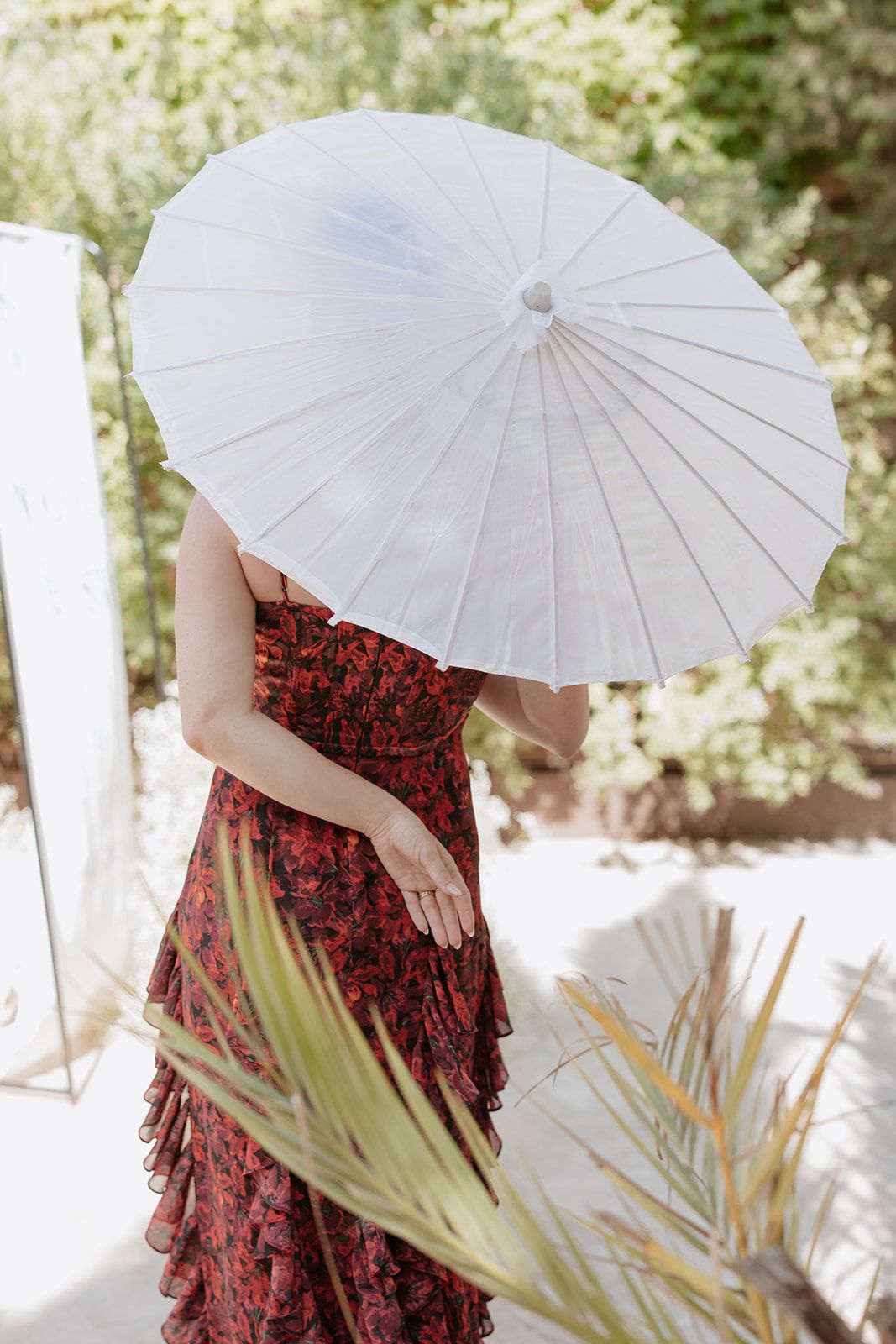 Person in red dress holding white parasol amidst tropical foliage in bright sunlight.