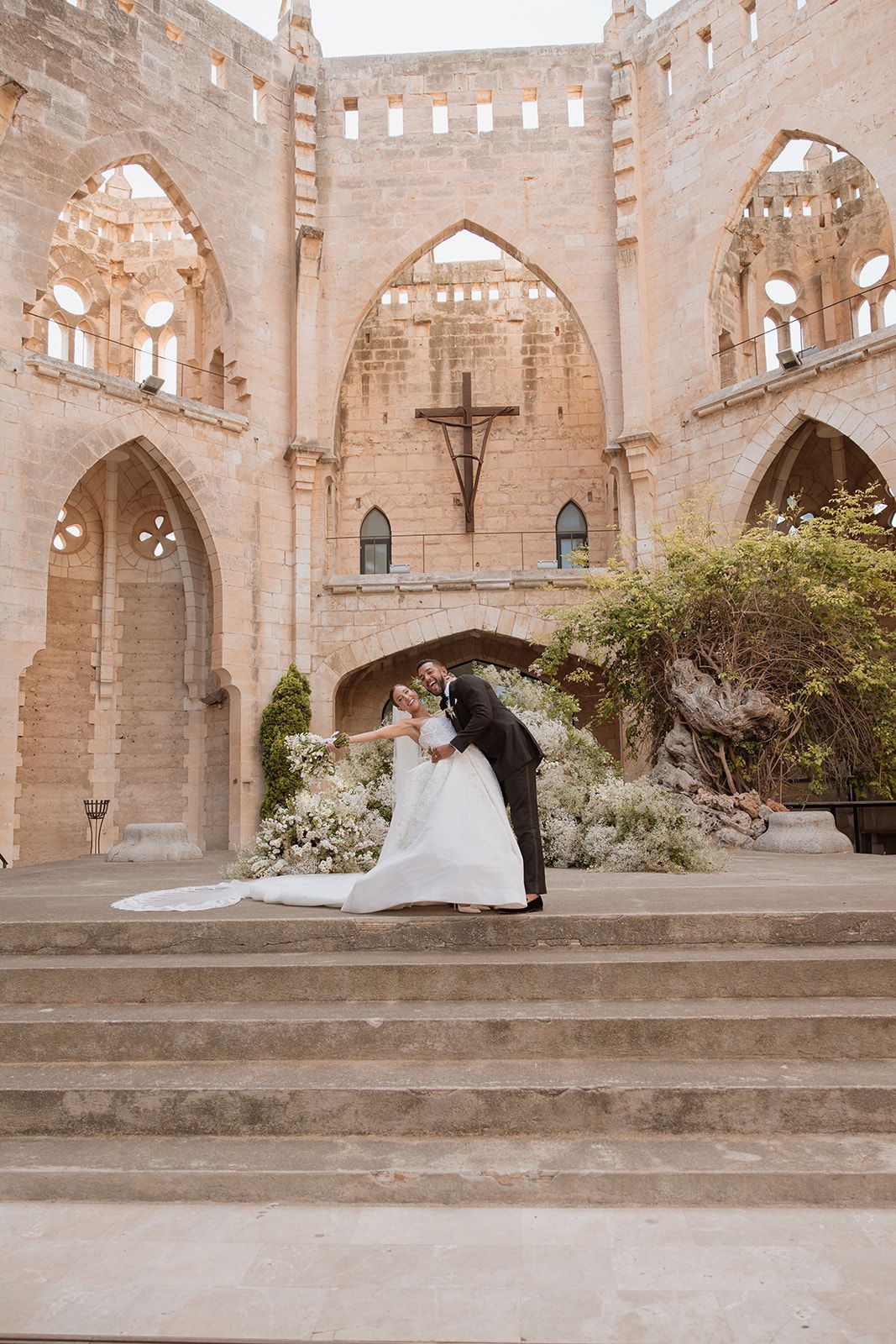 Golden hour wedding portrait on stone steps with gothic architecture and floral decorations.