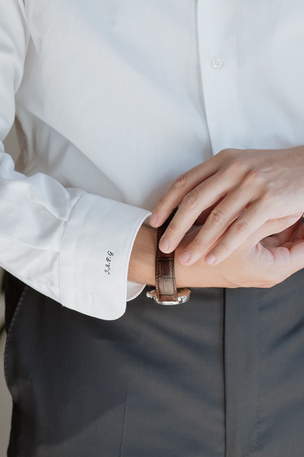 Close-up of hands adjusting a brown leather watch strap on wrist with white shirt sleeve.