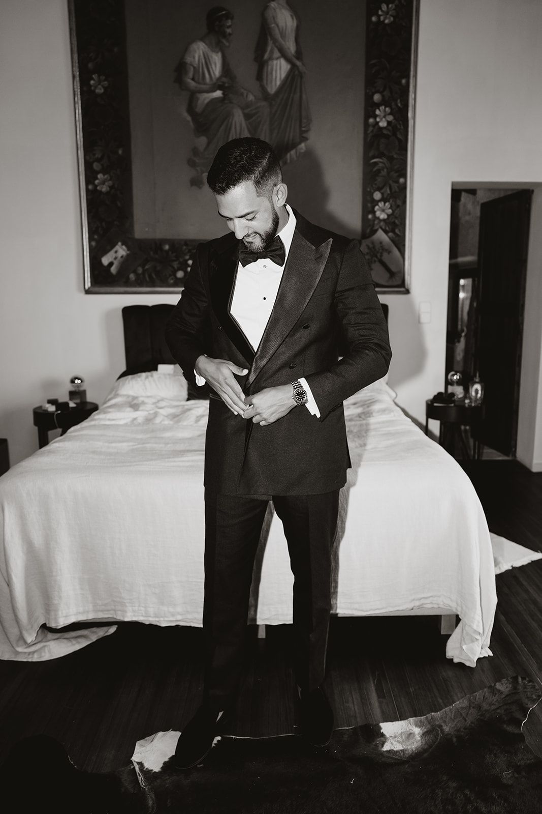 Black and white photo of someone in a tuxedo standing in an elegant bedroom.