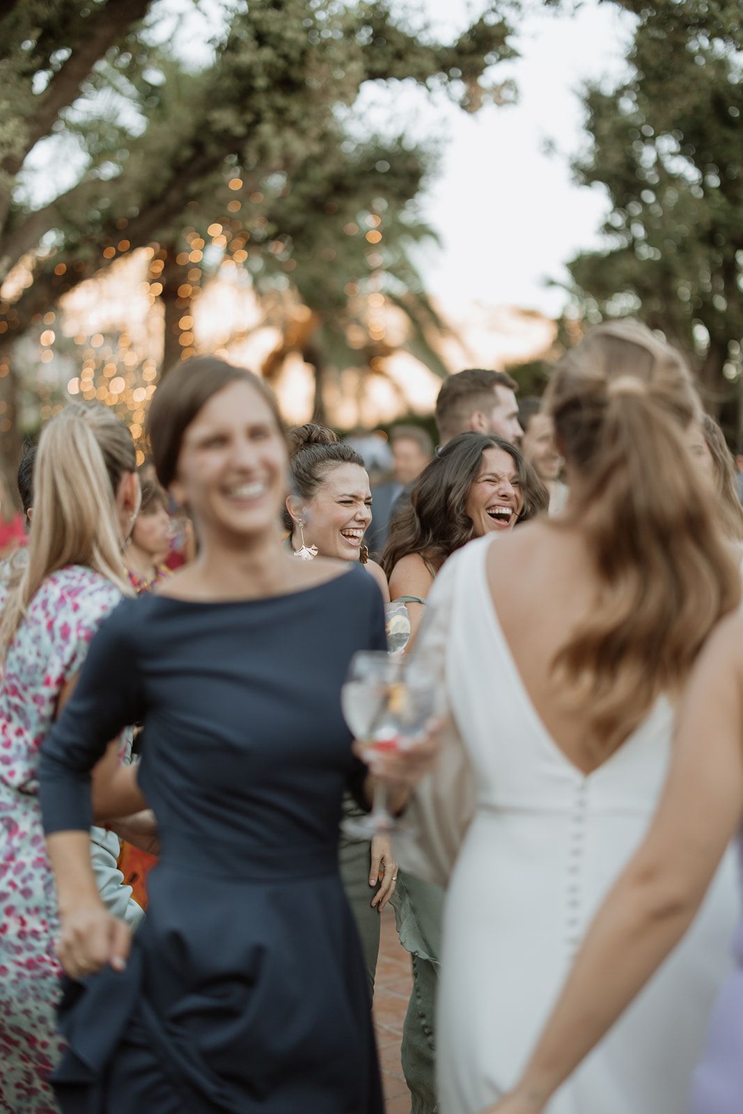 Wedding guests laugh together during sunset at an outdoor reception with string lights.