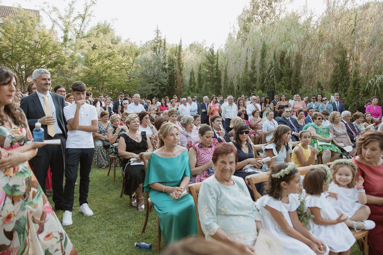 Wedding guests seated outdoors on white chairs during a garden ceremony.