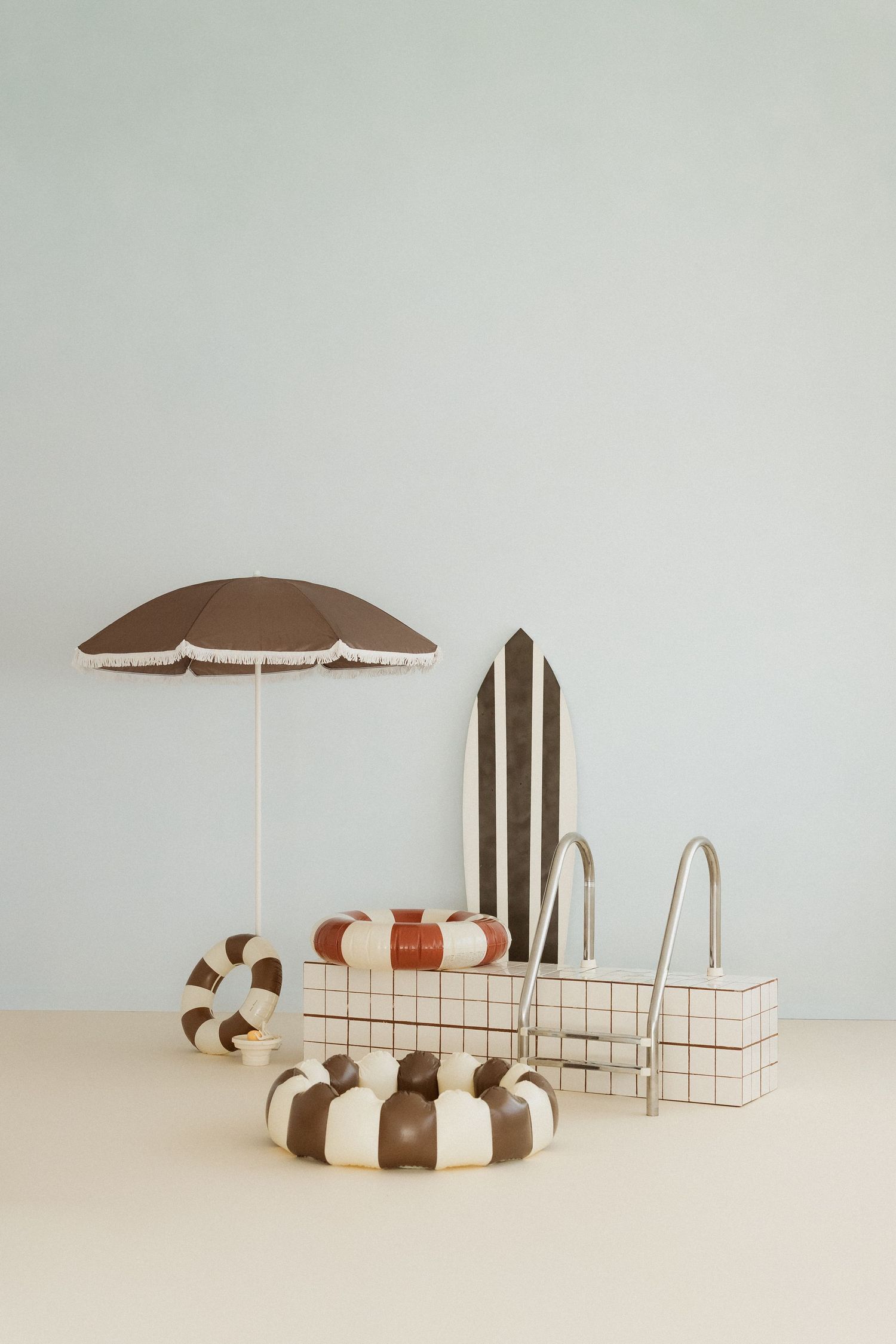 Retro-style miniature pool scene with striped umbrella, inflatable rings, surfboard, and tiled pool ladder.