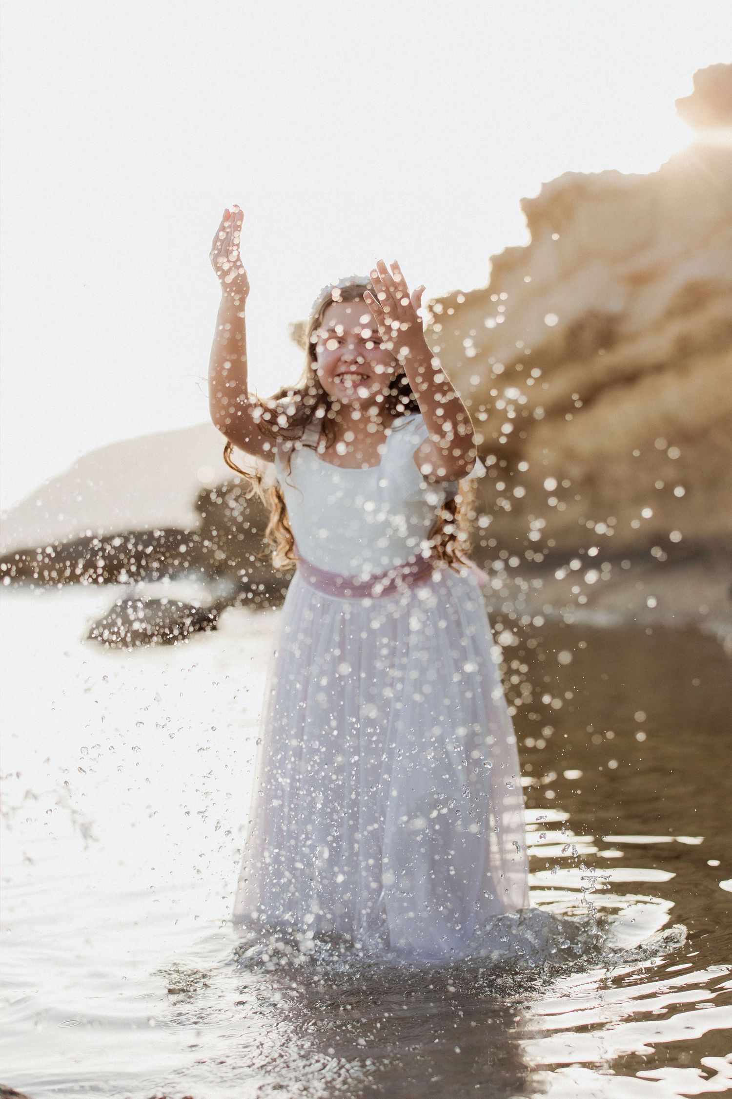 Young girl in white dress splashing water at beach, smiling joyfully with arms raised in golden sunlight.