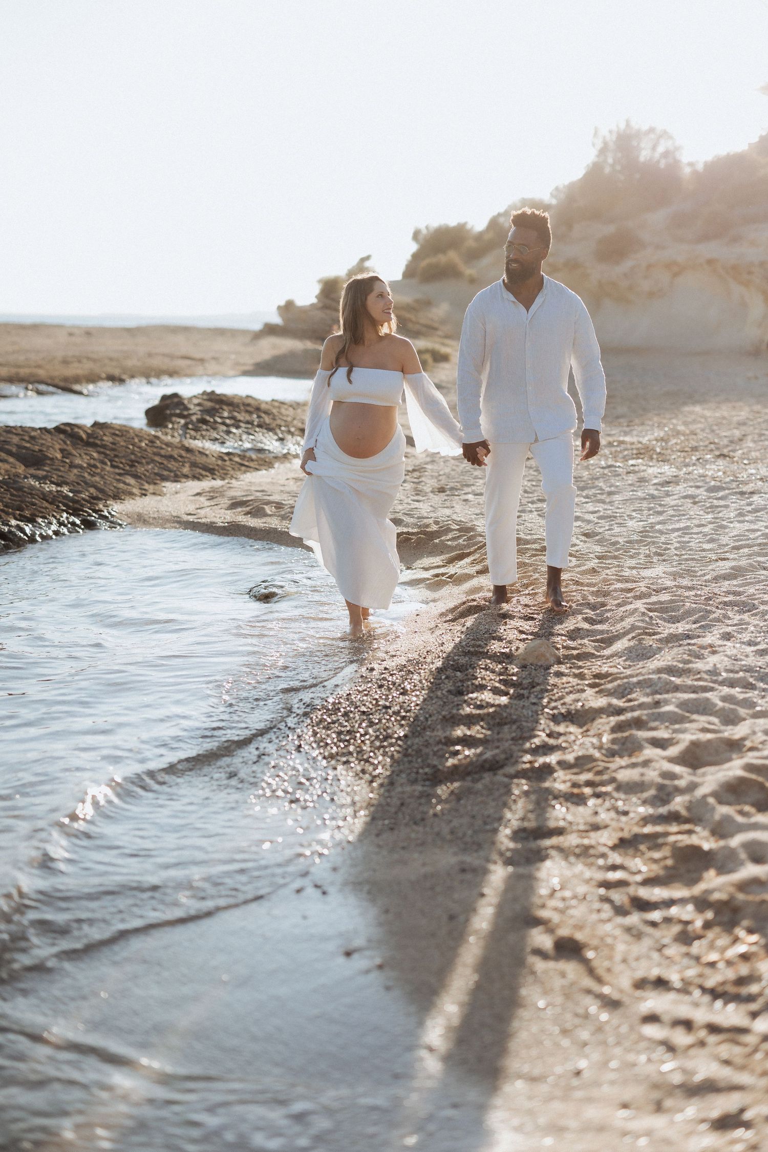 A couple walks along a sandy beach at sunset casting long shadows on the shoreline while wearing white outfits.