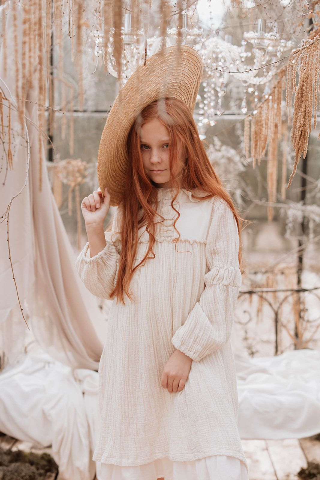 Elegant pose with wide-brim straw hat against crystal and fabric backdrop.