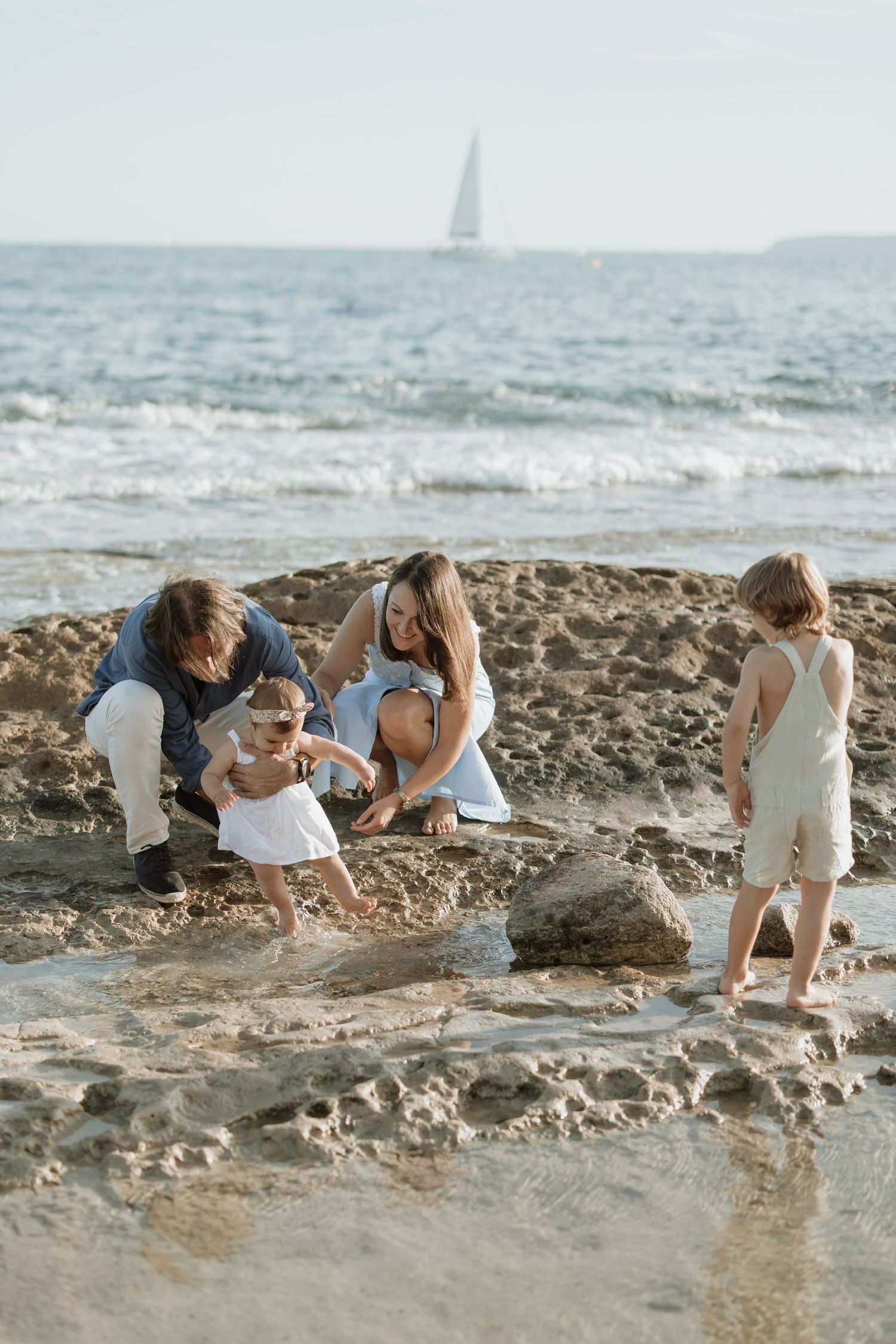 Family exploring rocky beach together while sailboat sails in distance.