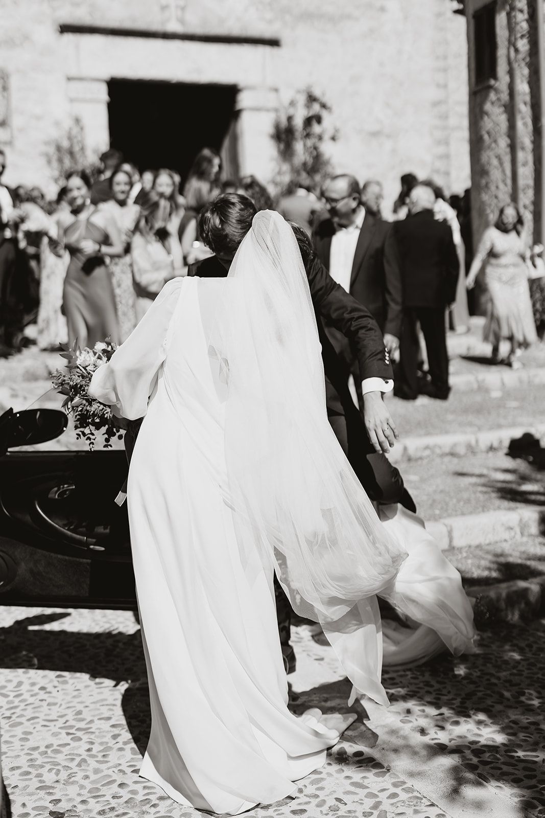 A bride in a flowing white dress with a long veil exits a building while wedding guests look on.