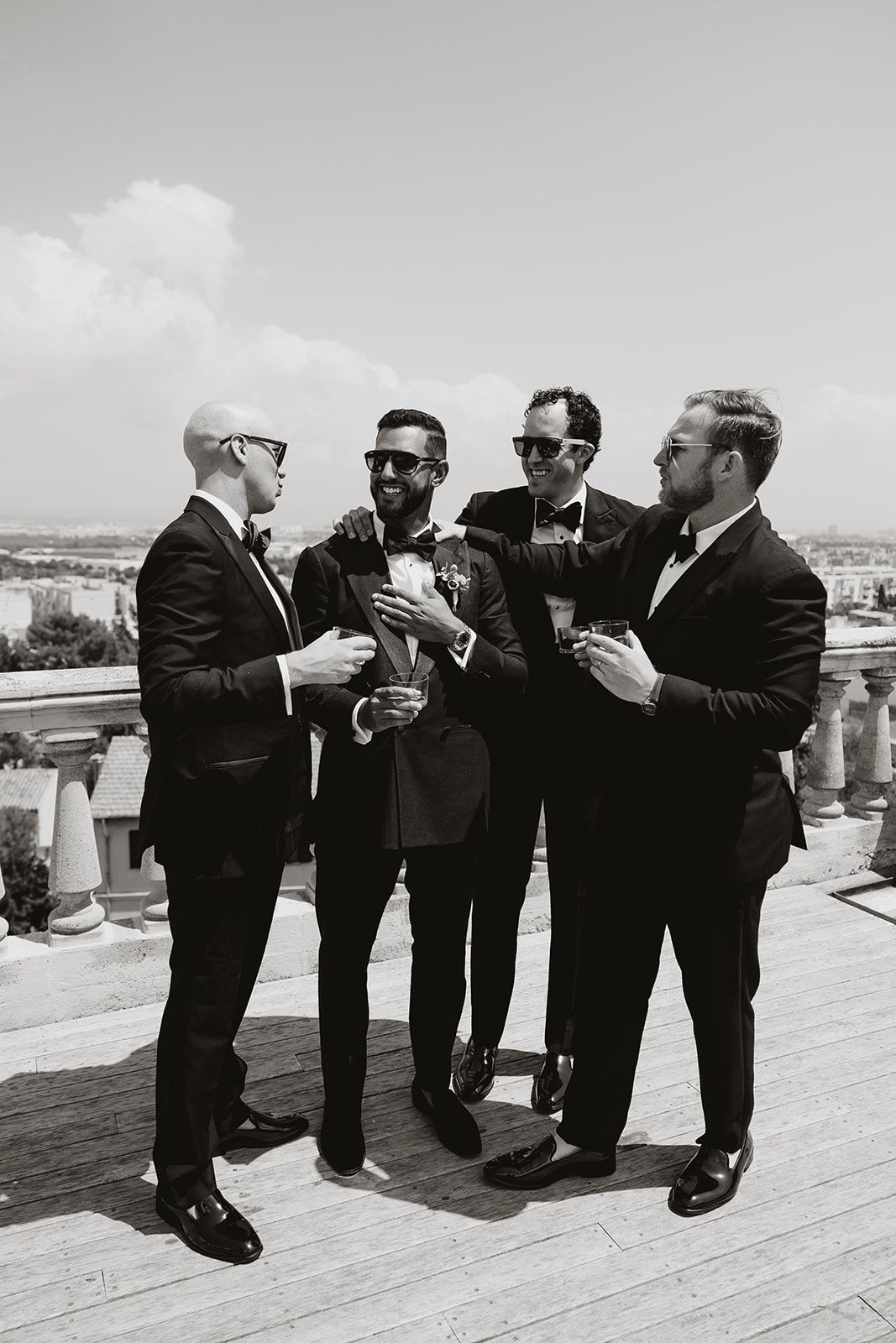Four groomsmen in black suits and sunglasses share drinks and laughs on an outdoor terrace.