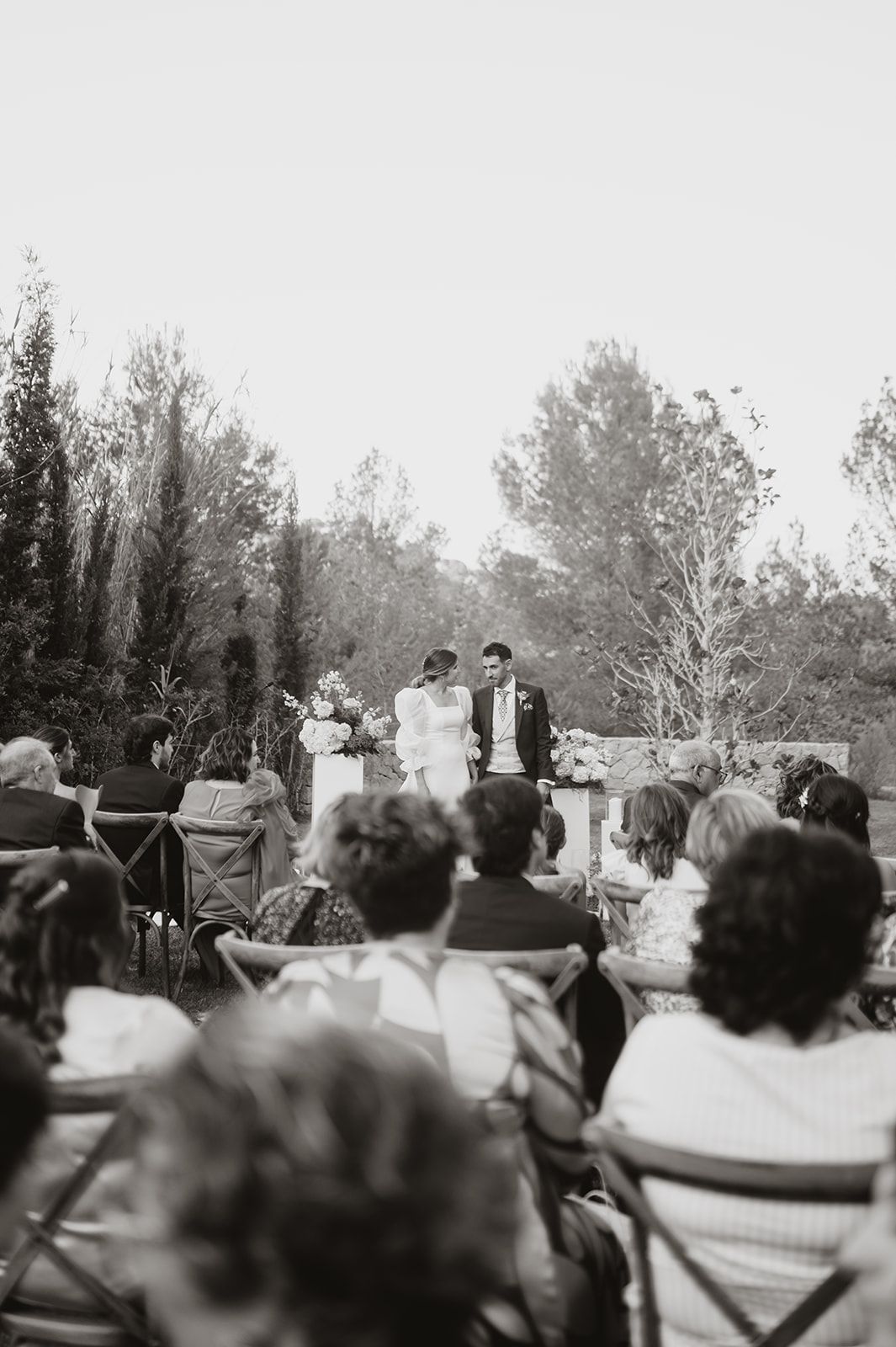 A black and white photo of an outdoor wedding ceremony with guests seated on lawn chairs.