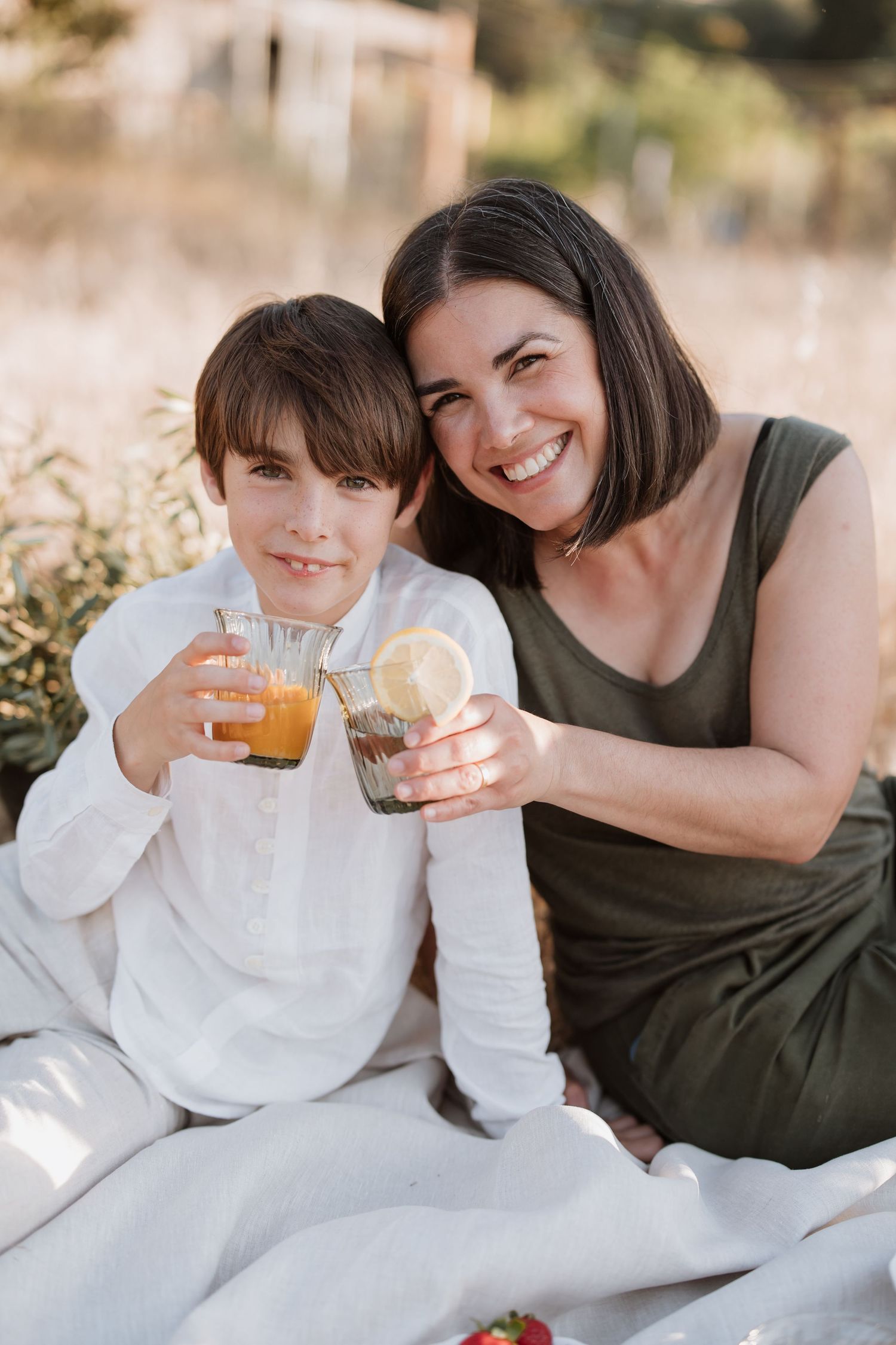 Two people enjoying refreshing drinks while sitting on a white blanket for an outdoor picnic on a sunny day.