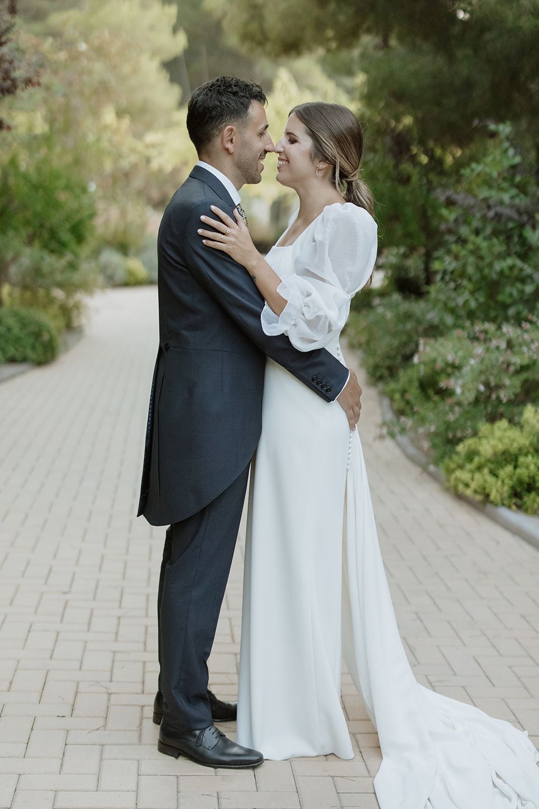 Bride in elegant white dress with flowing sleeves embraces groom in formal tailcoat on garden path.