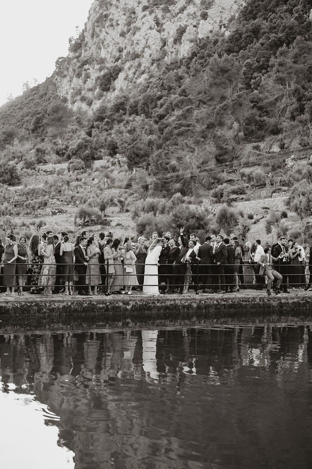 Wedding party and guests lined up along waterfront with mountain backdrop reflected in water.