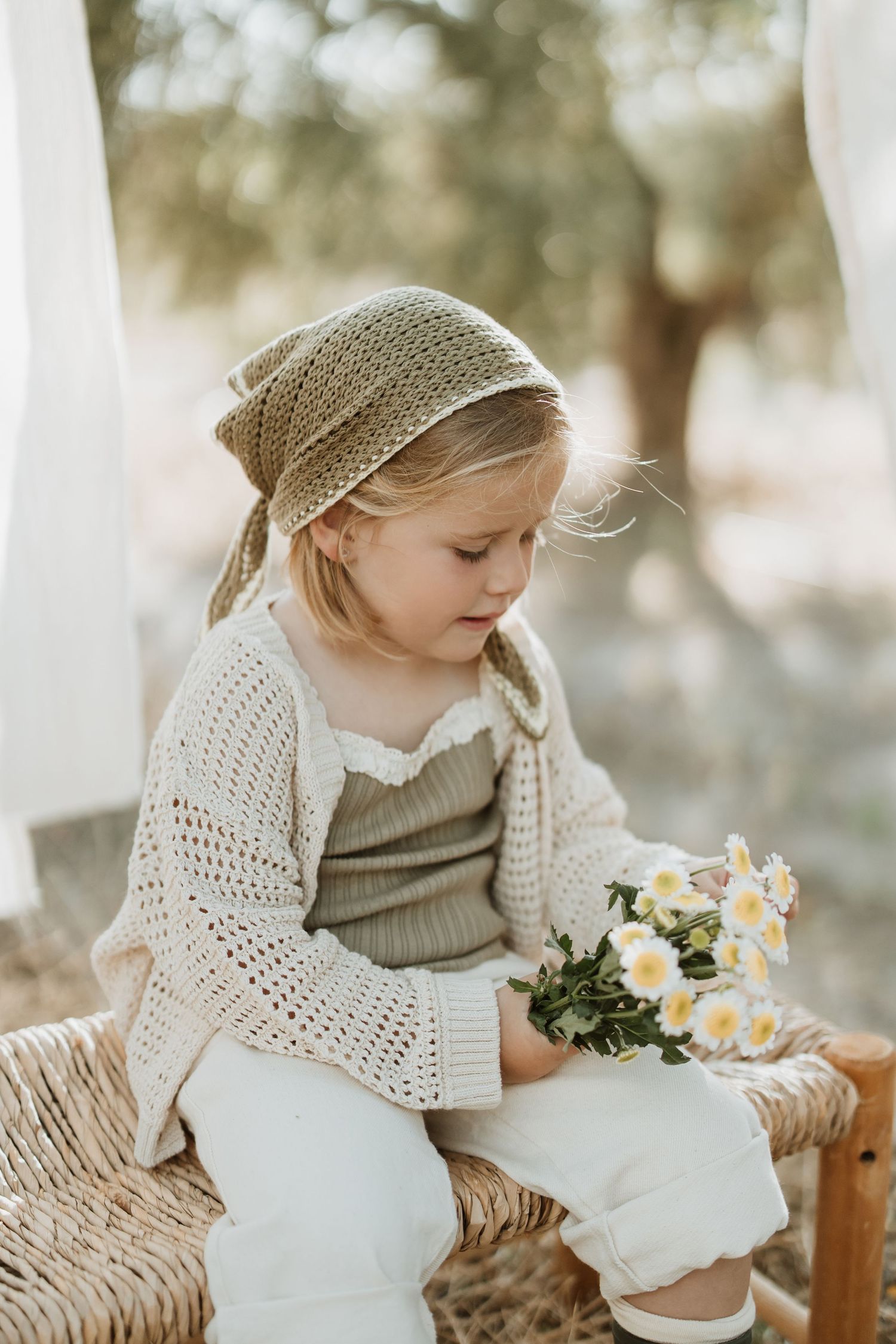 Child in headscarf and knit cardigan looking at flowers while sitting on woven bench.