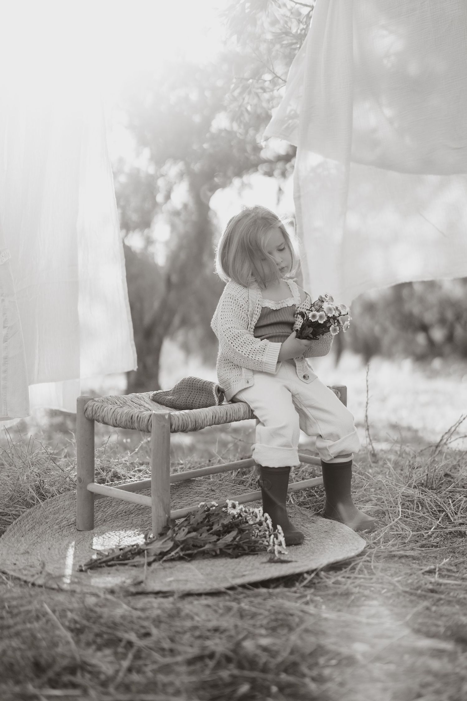 Black and white photo of child on rustic stool holding flowers outdoors.
