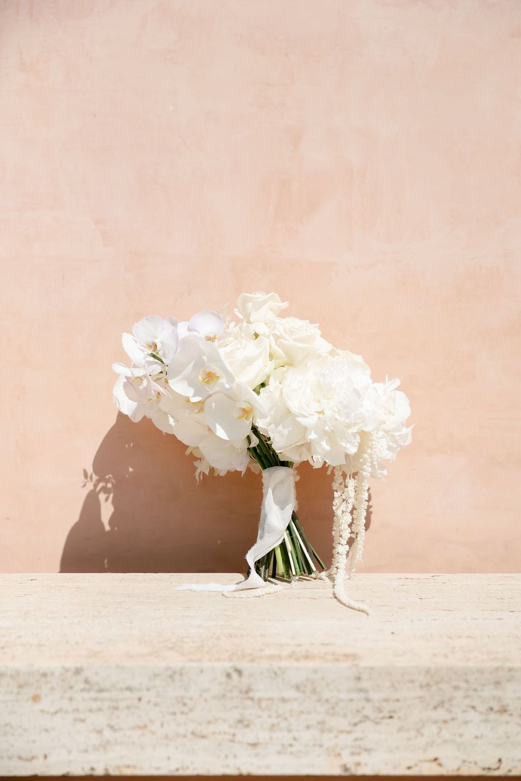 White peony bridal bouquet against peachy pink wall with dramatic shadows.