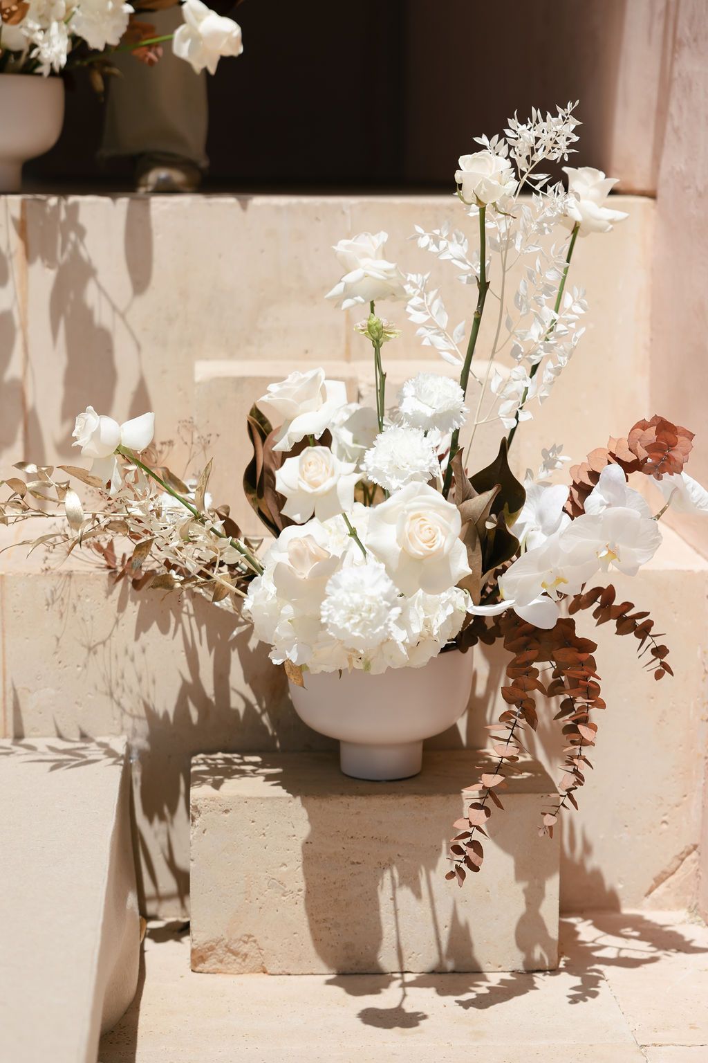 White floral arrangement with roses and orchids in a modern vase casting dramatic shadows.