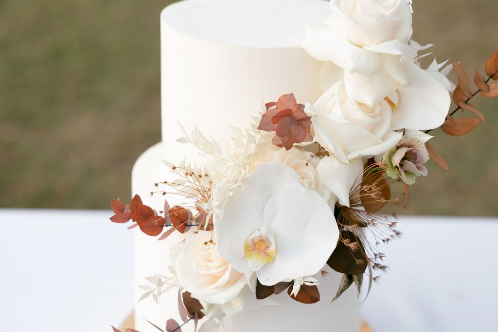 White tiered wedding cake adorned with white orchids and copper-colored botanical elements.