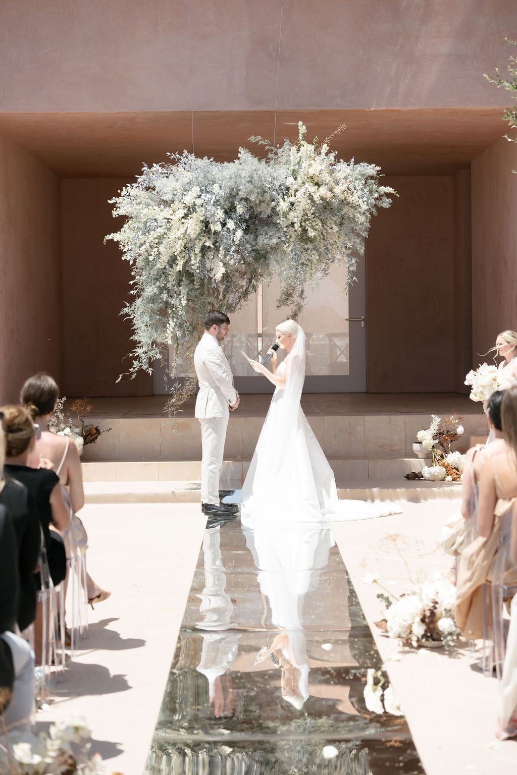 Wedding ceremony under a white floral arch with reflective aisle and guests seated on both sides.