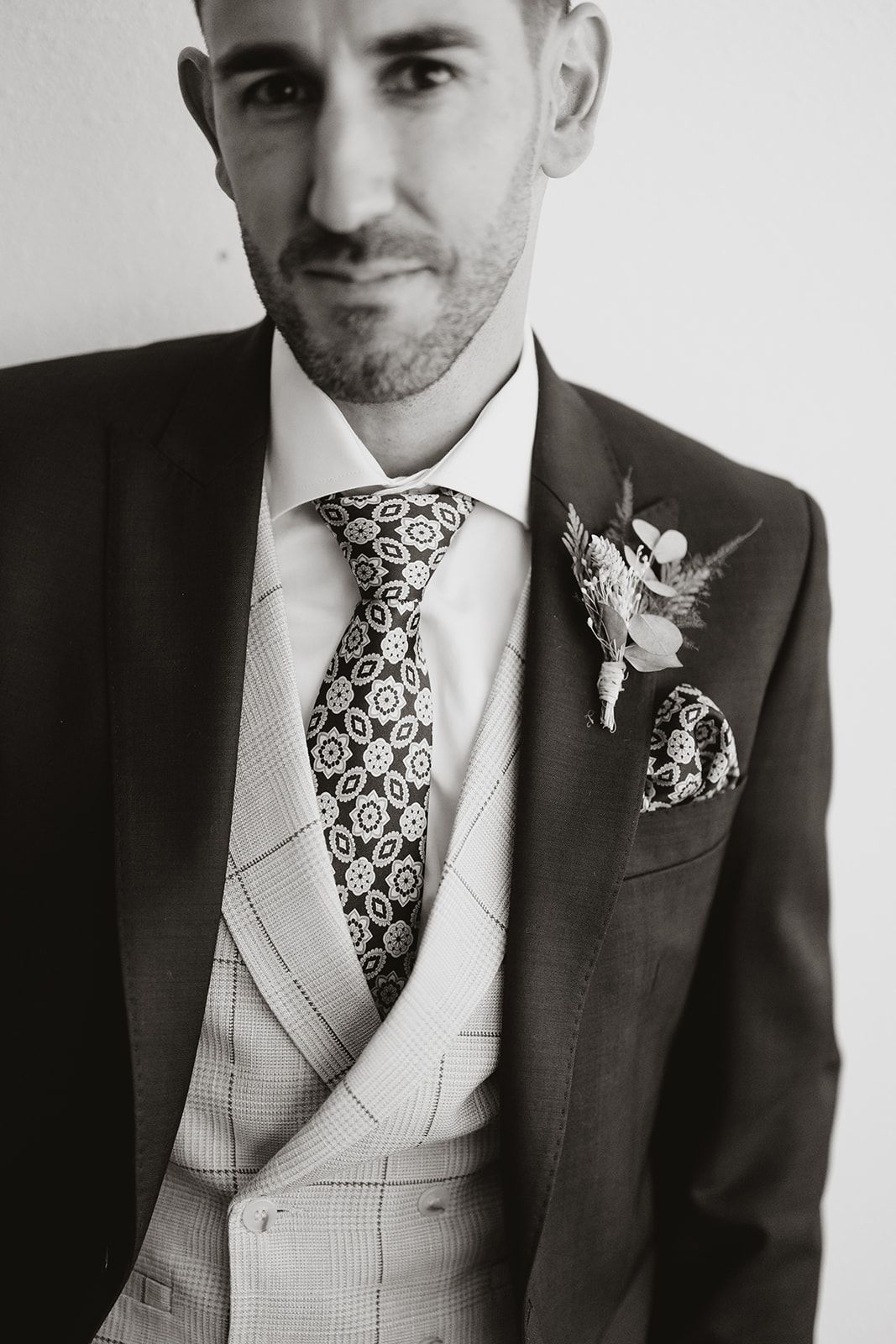 A detailed black and white close-up of a groom's attire featuring a patterned tie and boutonniere.