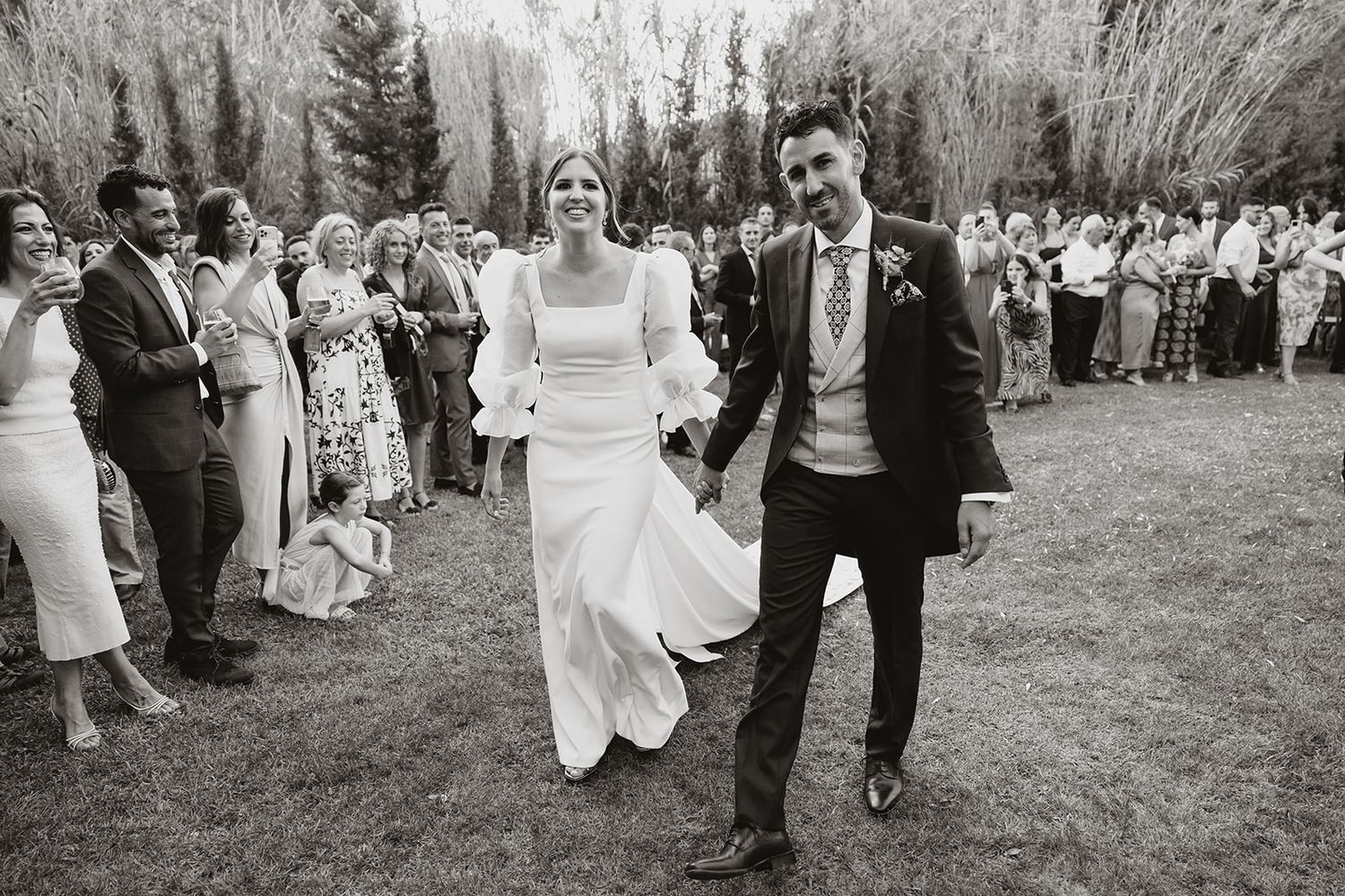 Black and white photo captures joyful newlyweds walking through crowd of wedding guests.