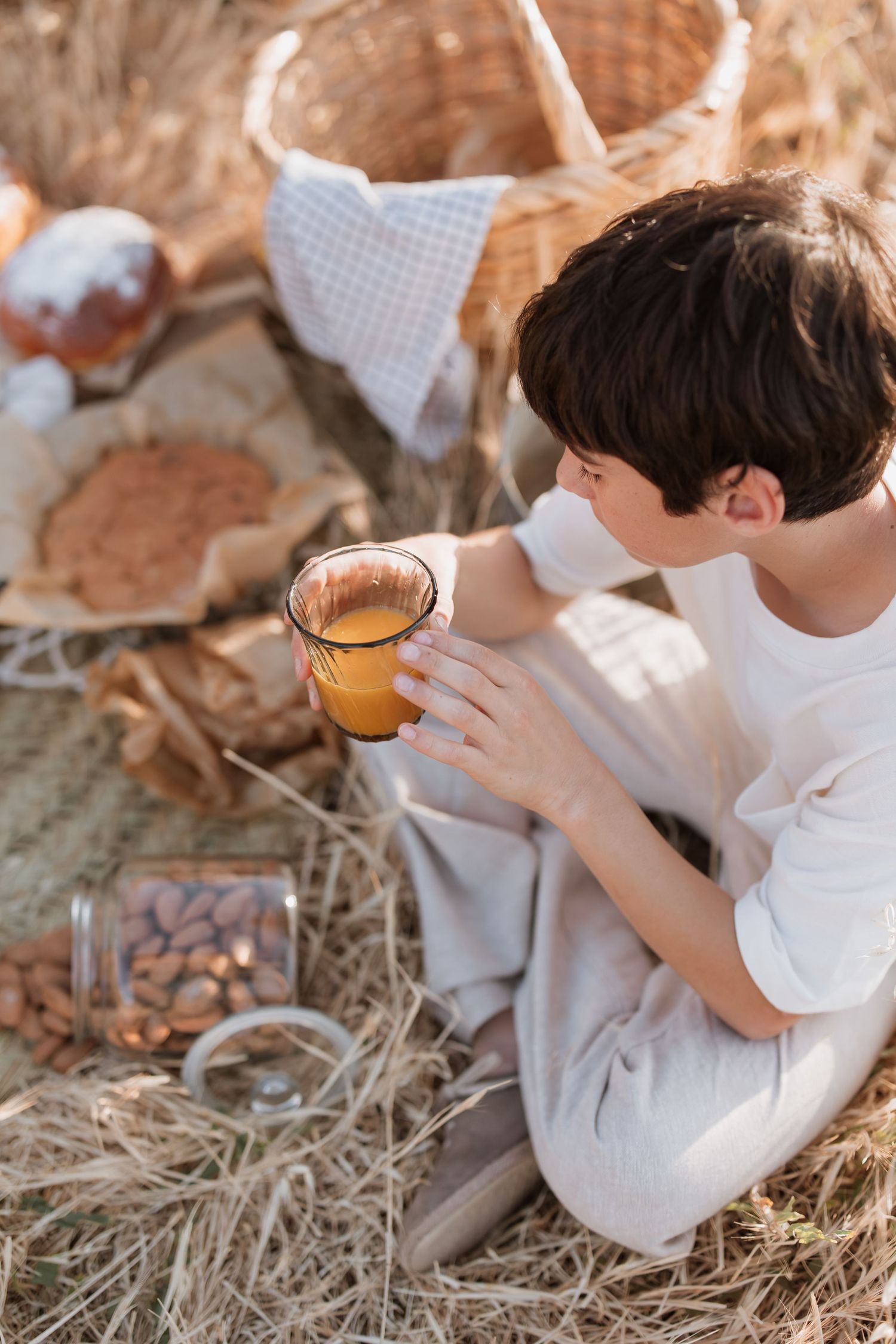 Child in white clothing sitting on hay and drinking a golden beverage from a glass during an outdoor picnic scene.