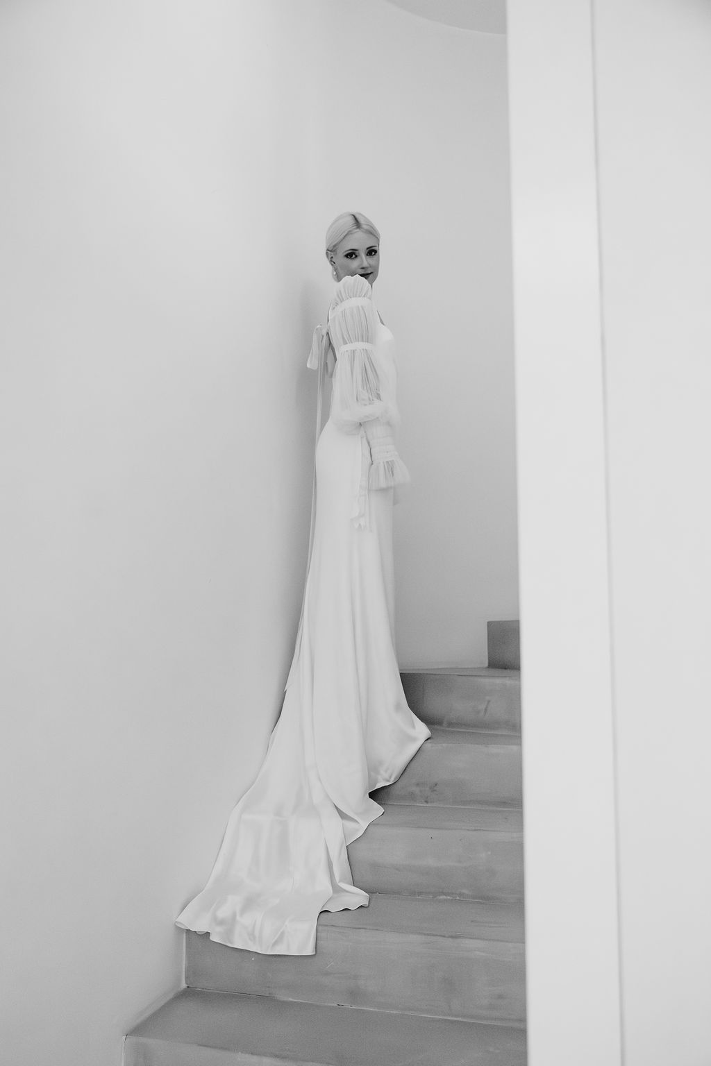A dramatic black and white photograph of a wedding dress on stairs against stark white walls.