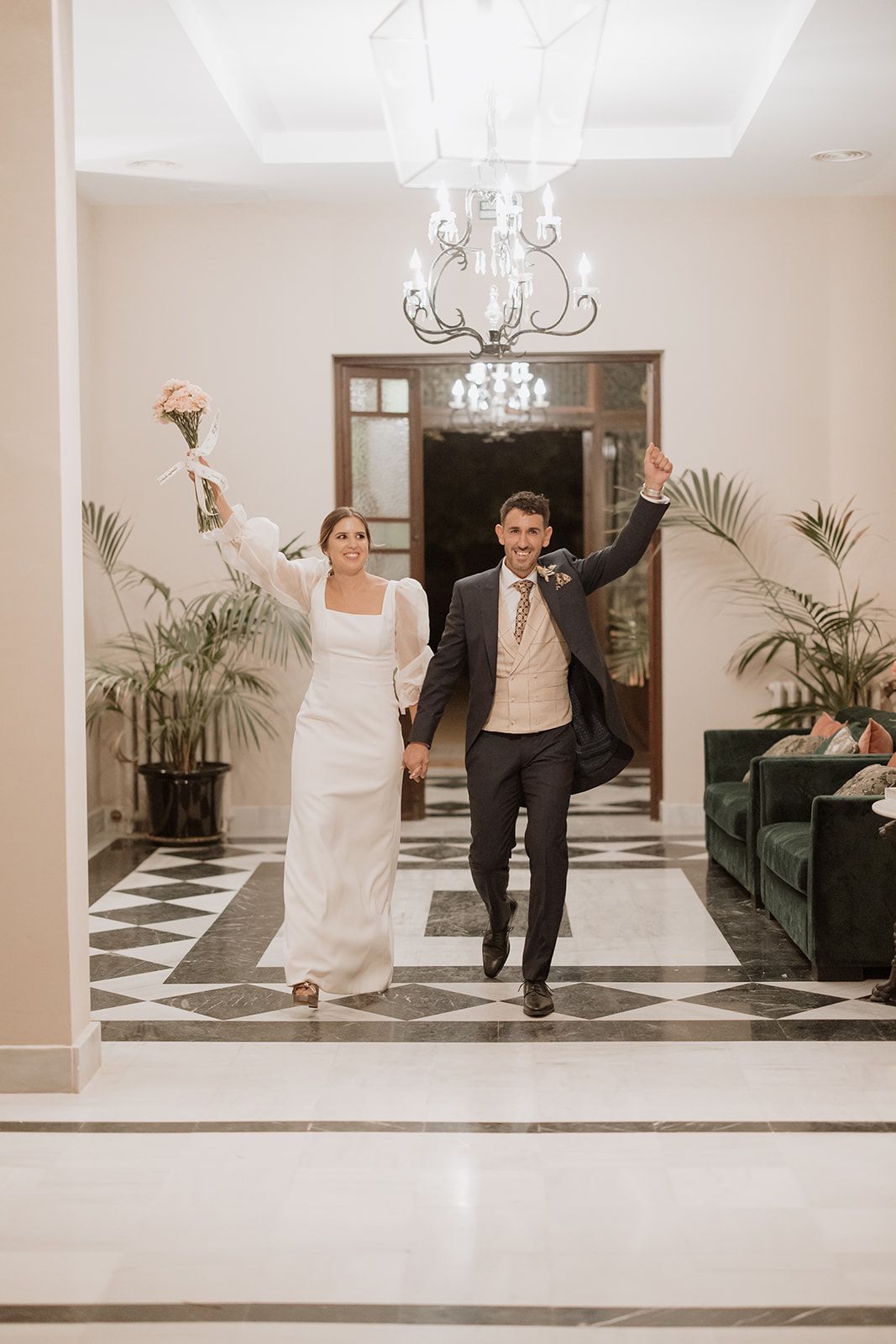 Newlyweds make joyful entrance into reception hall with elegant chandelier and checkered floor.