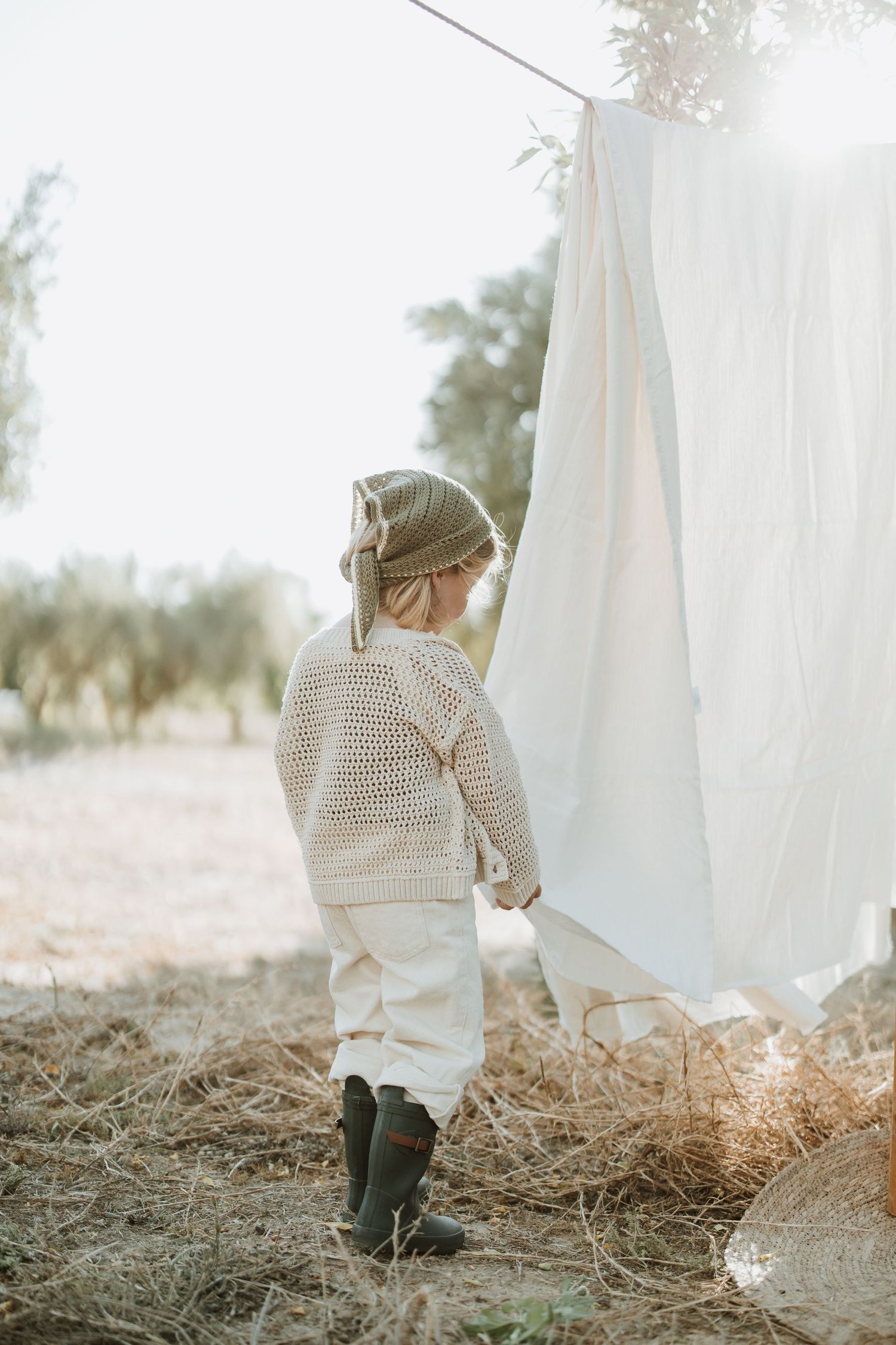 Child in boots and sweater stands by white sheets drying on clothesline at sunset.