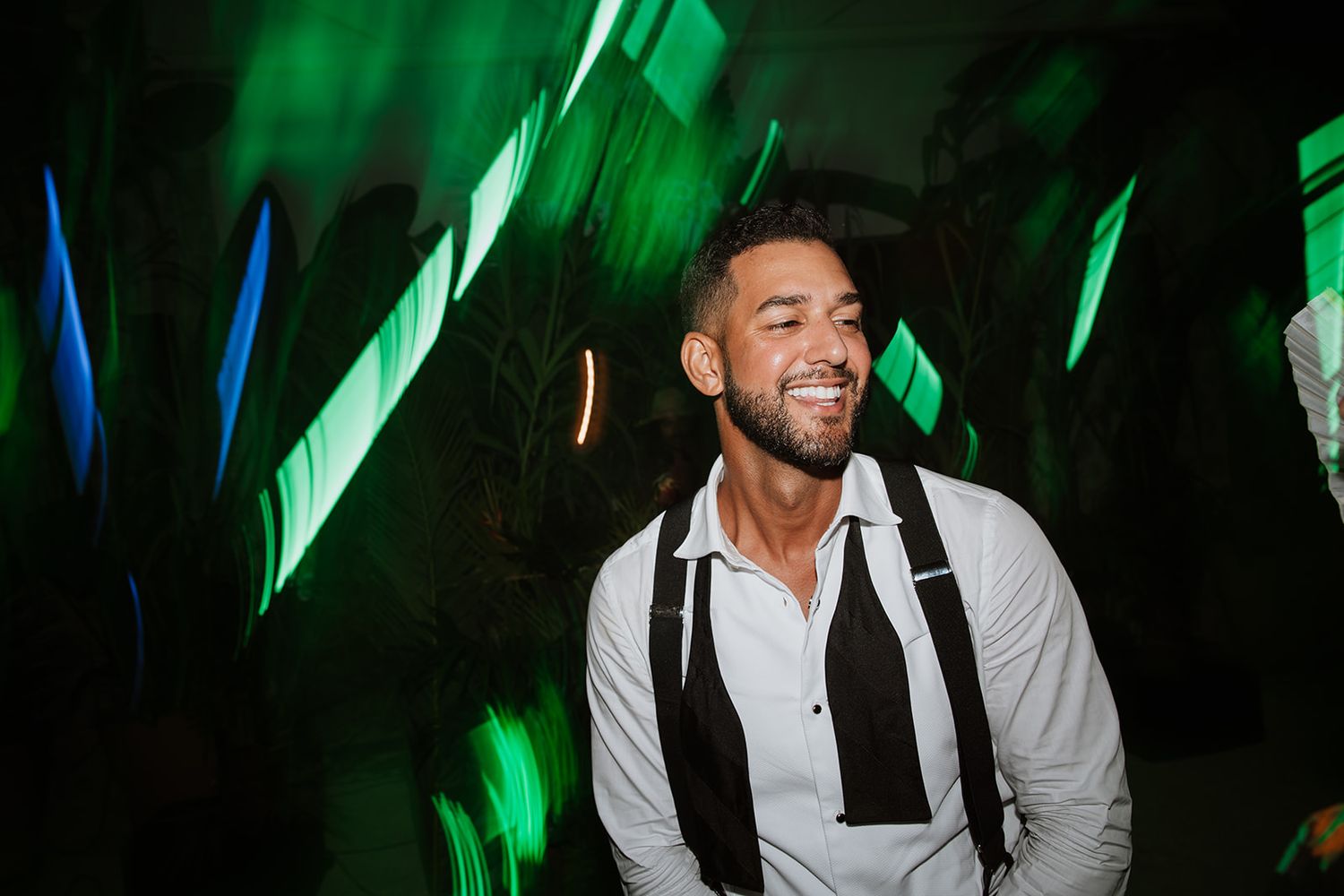 Person in white shirt and suspenders smiling joyfully under colored party lights.