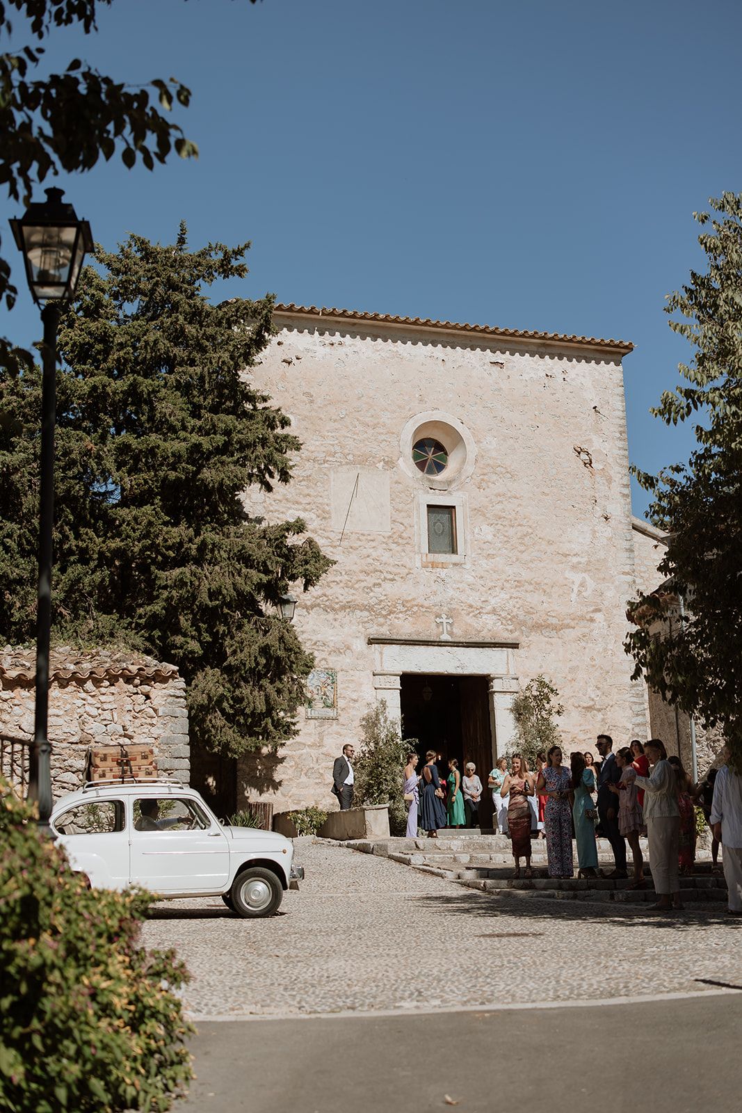 Vintage white car parked in front of historic stone church with gathering crowd.