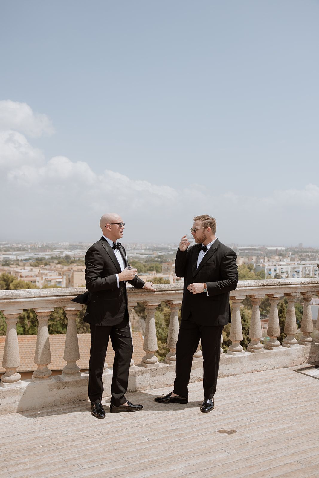 Two people in tuxedos share conversation on balcony overlooking city landscape.