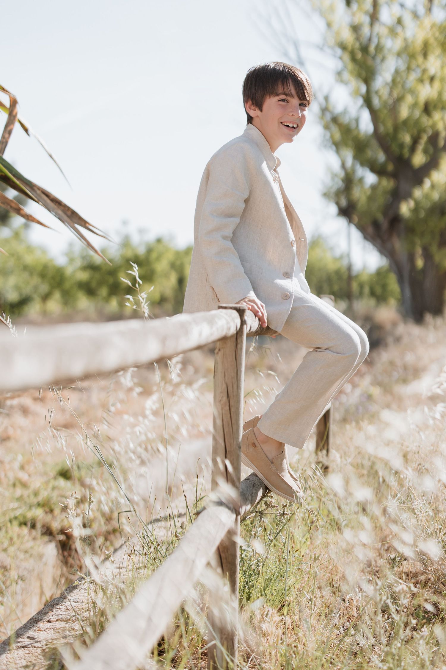 A person in a beige suit sits casually on a wooden fence in a sunlit field with dry grass and trees in background.