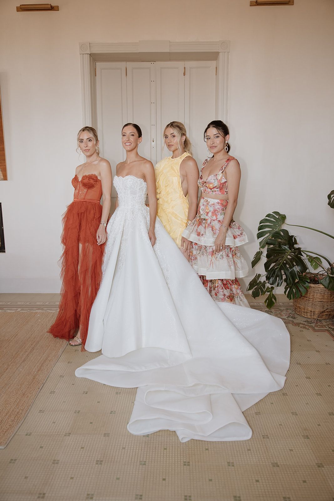 Wedding party in colorful dresses gathered in a bright room with white walls.